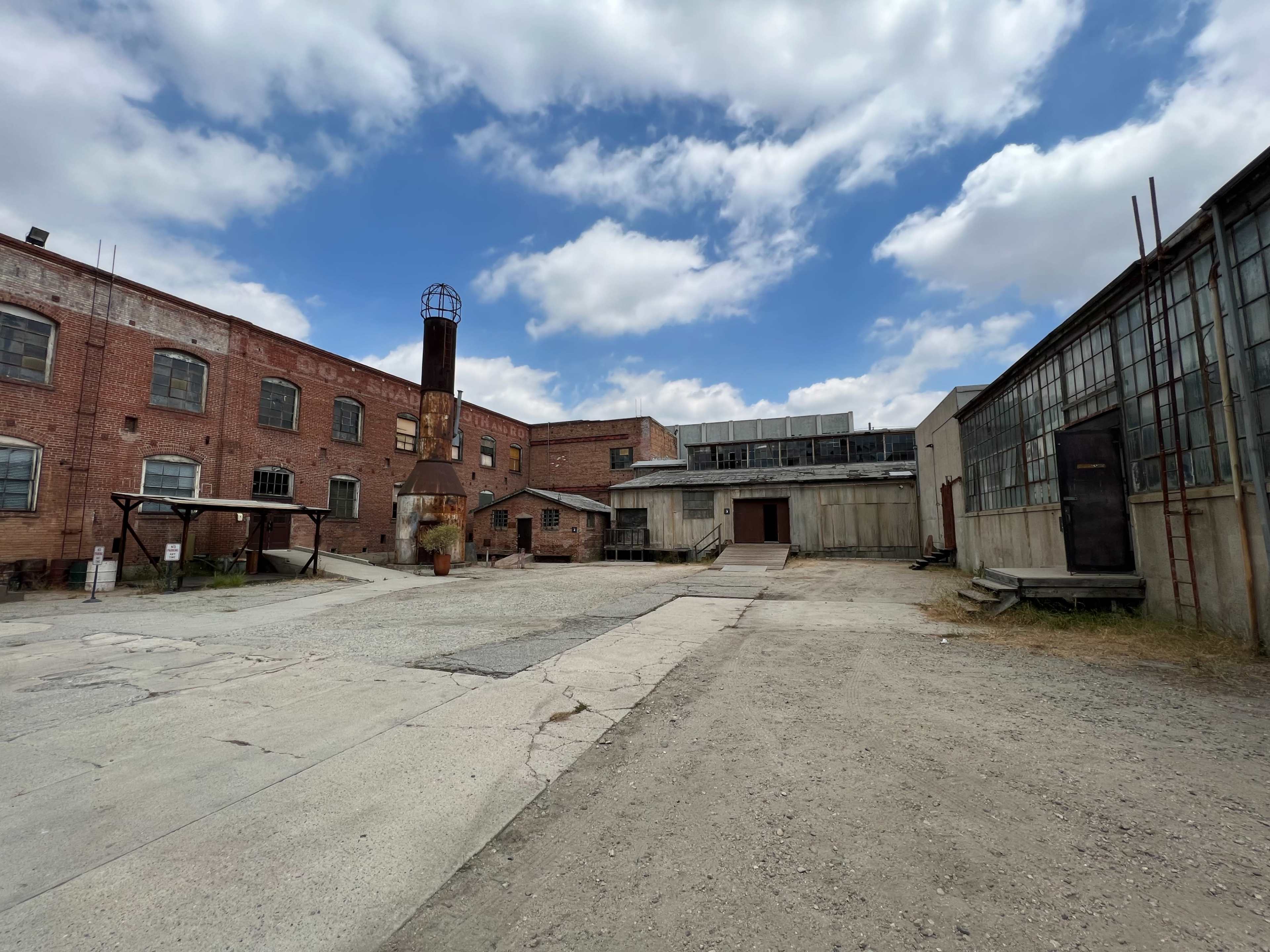 A deserted industrial area with brick buildings, a smokestack, and a gravel pathway under a cloudy sky.