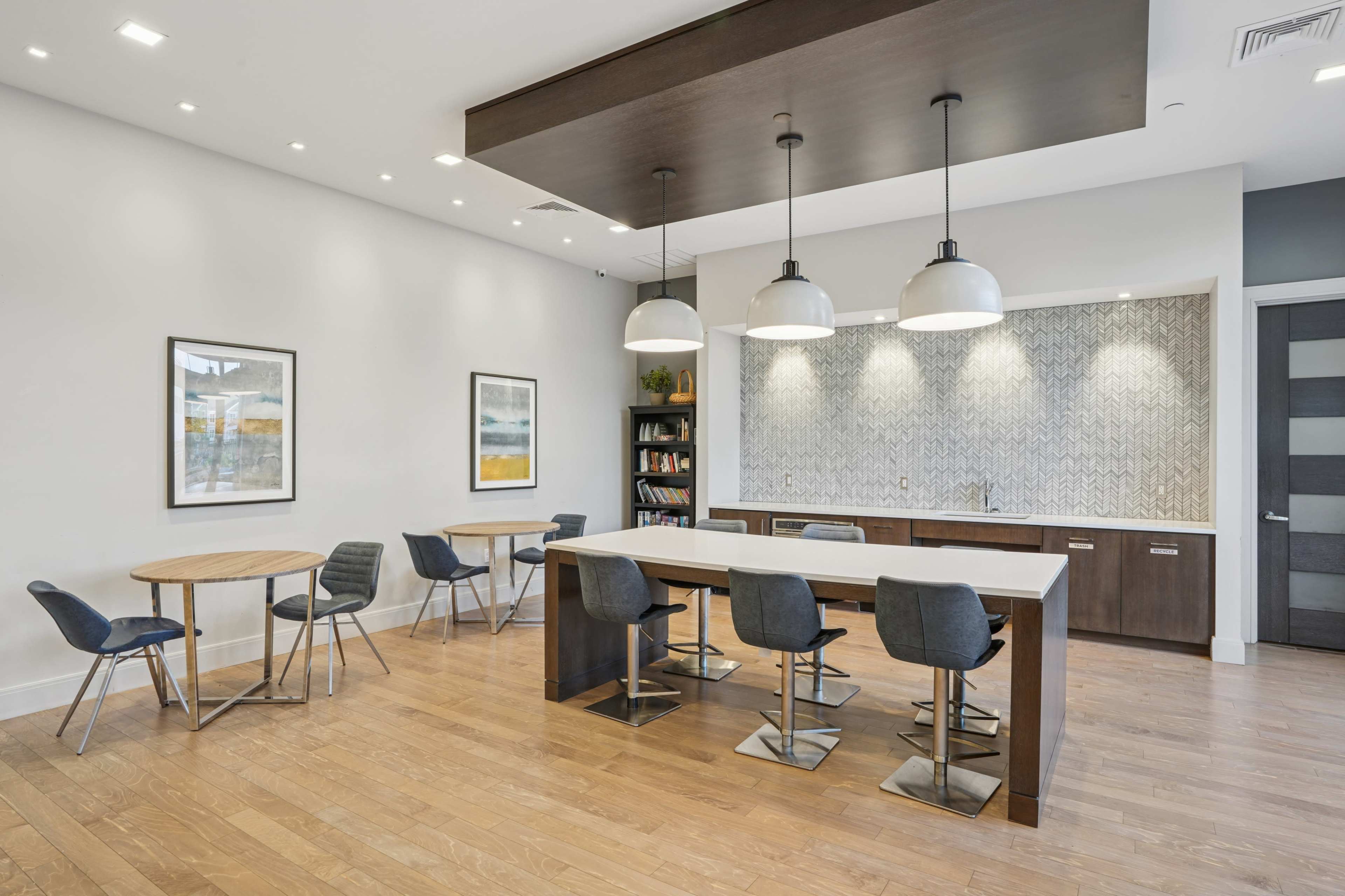 The image depicts a modern communal space featuring a large island bar with high stools, two round tables, and framed artwork on the walls.