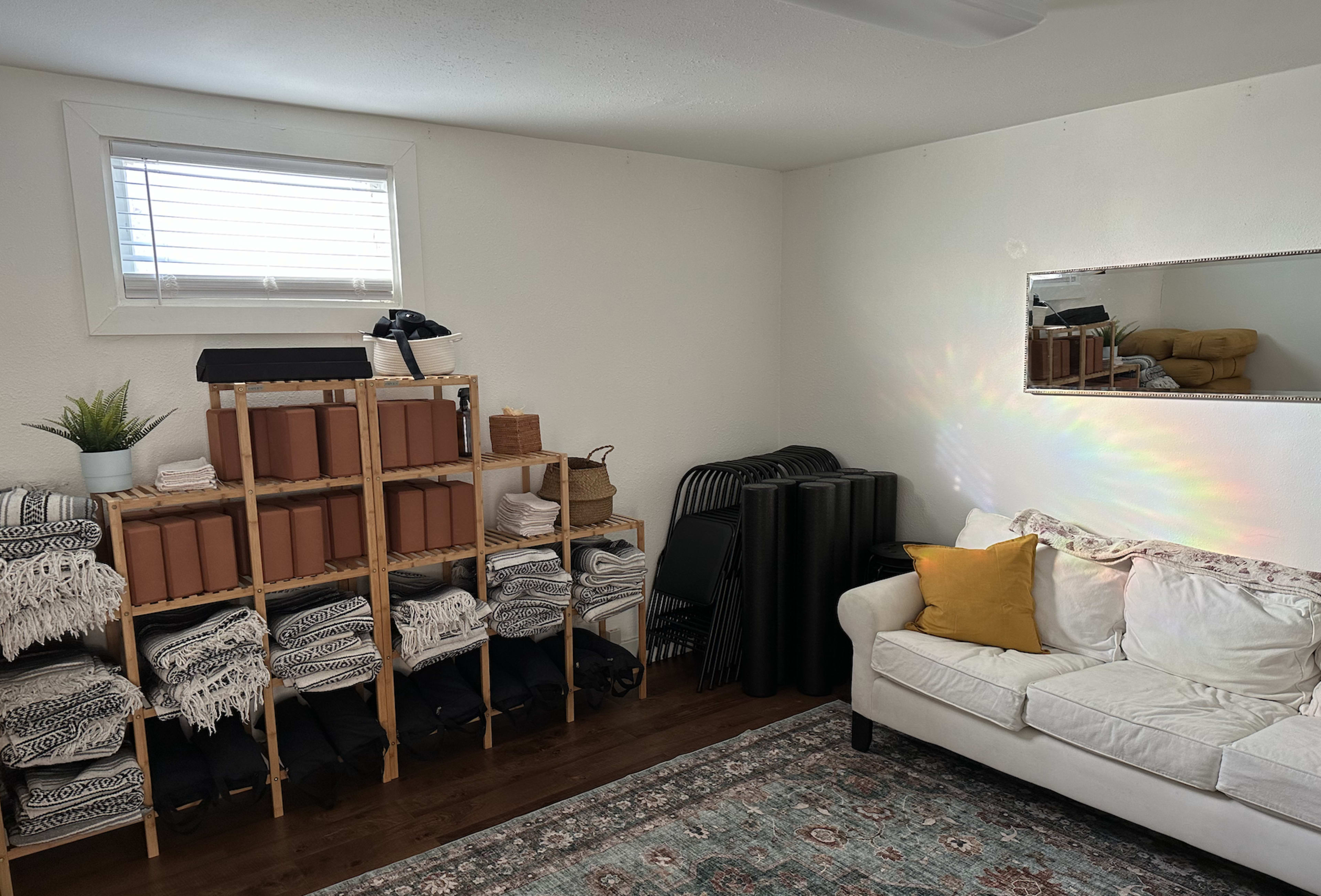 The image shows a neatly organized room with a shelf of stacked blankets and storage boxes, a couch with a yellow pillow, and a small rug on the floor.