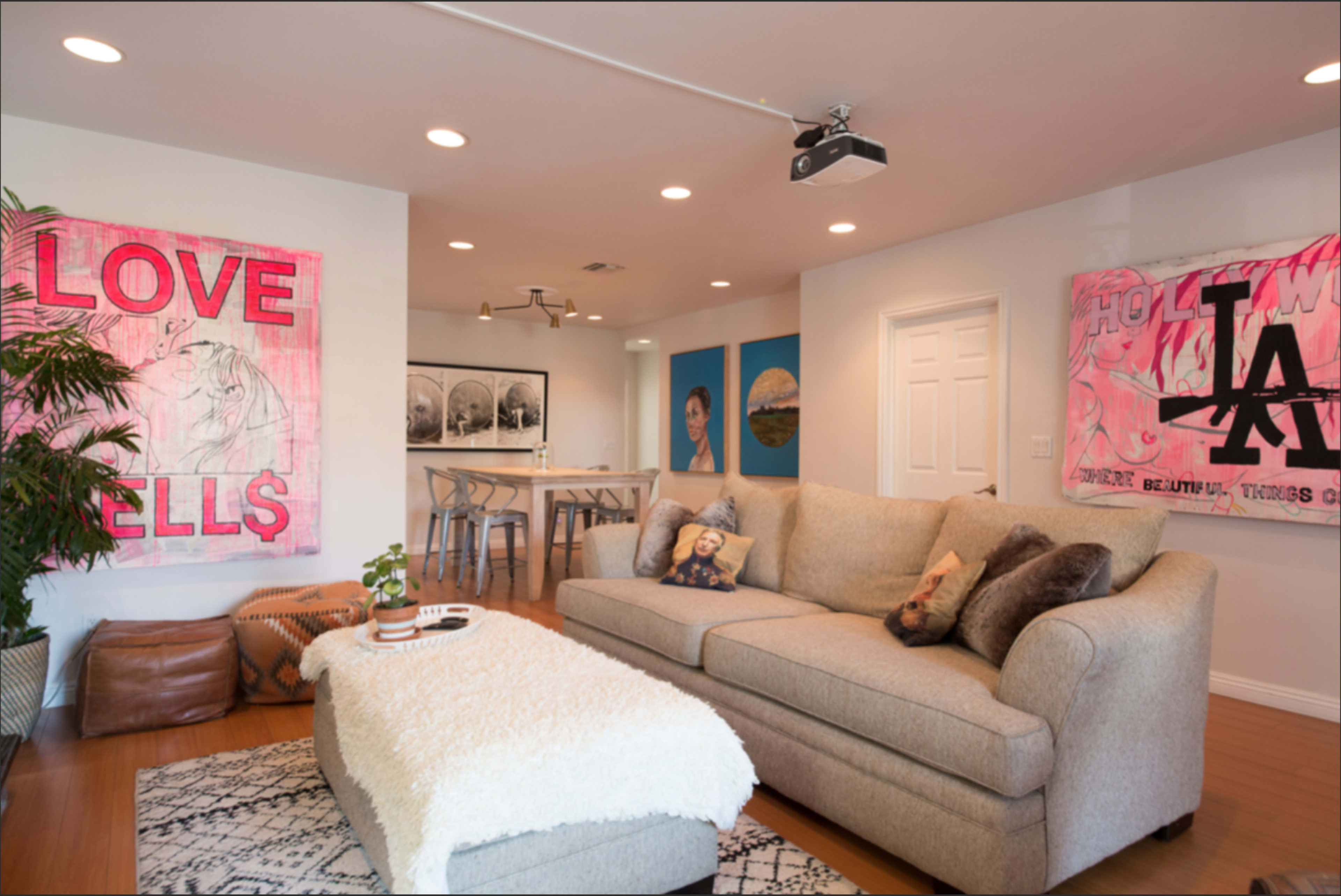 The living room features a beige sofa, a decorative rug, and colorful artwork on the walls, with a kitchen area visible in the background.