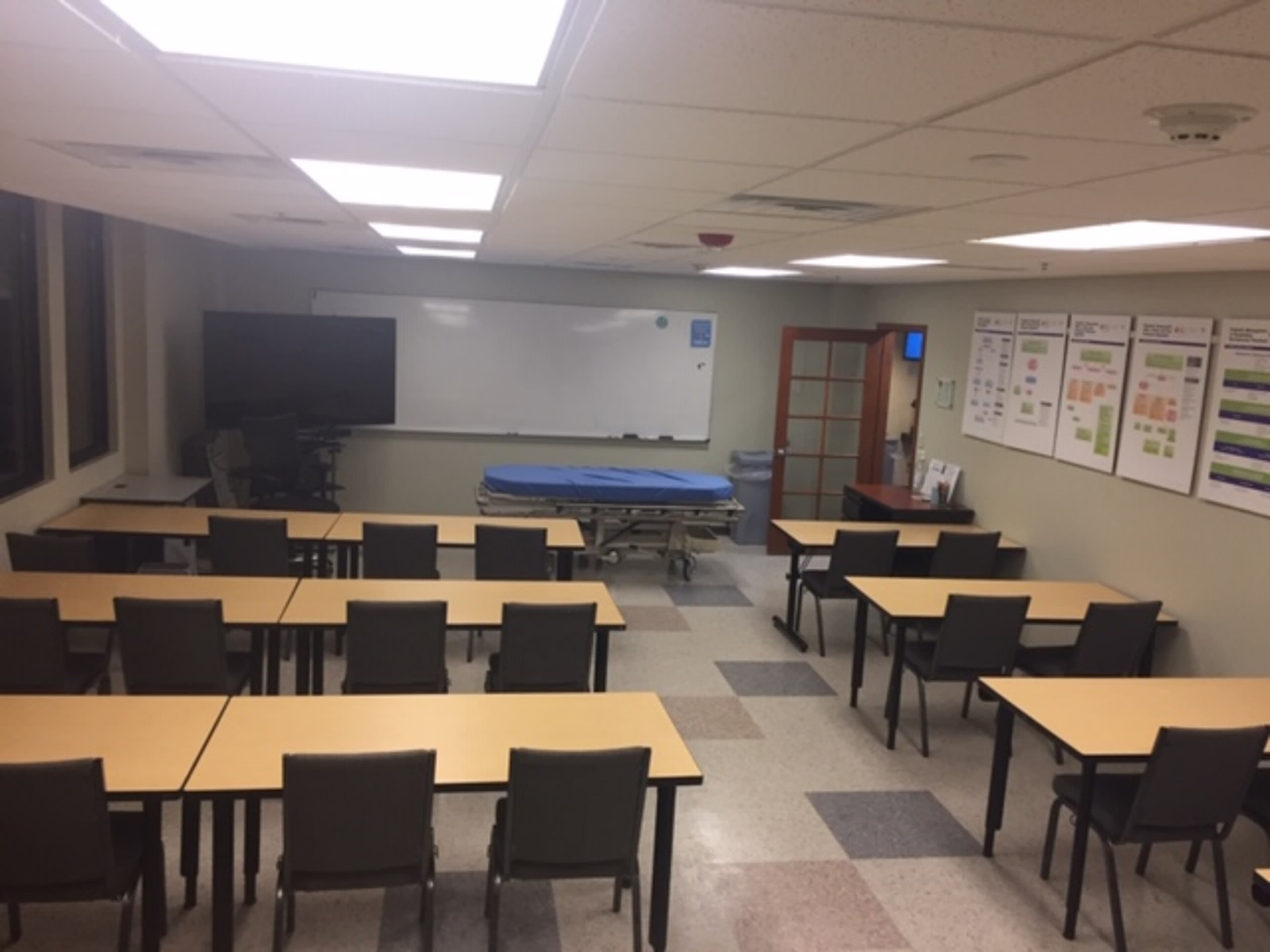 An empty classroom with rows of tables and chairs, featuring a whiteboard and a hospital bed at the front.