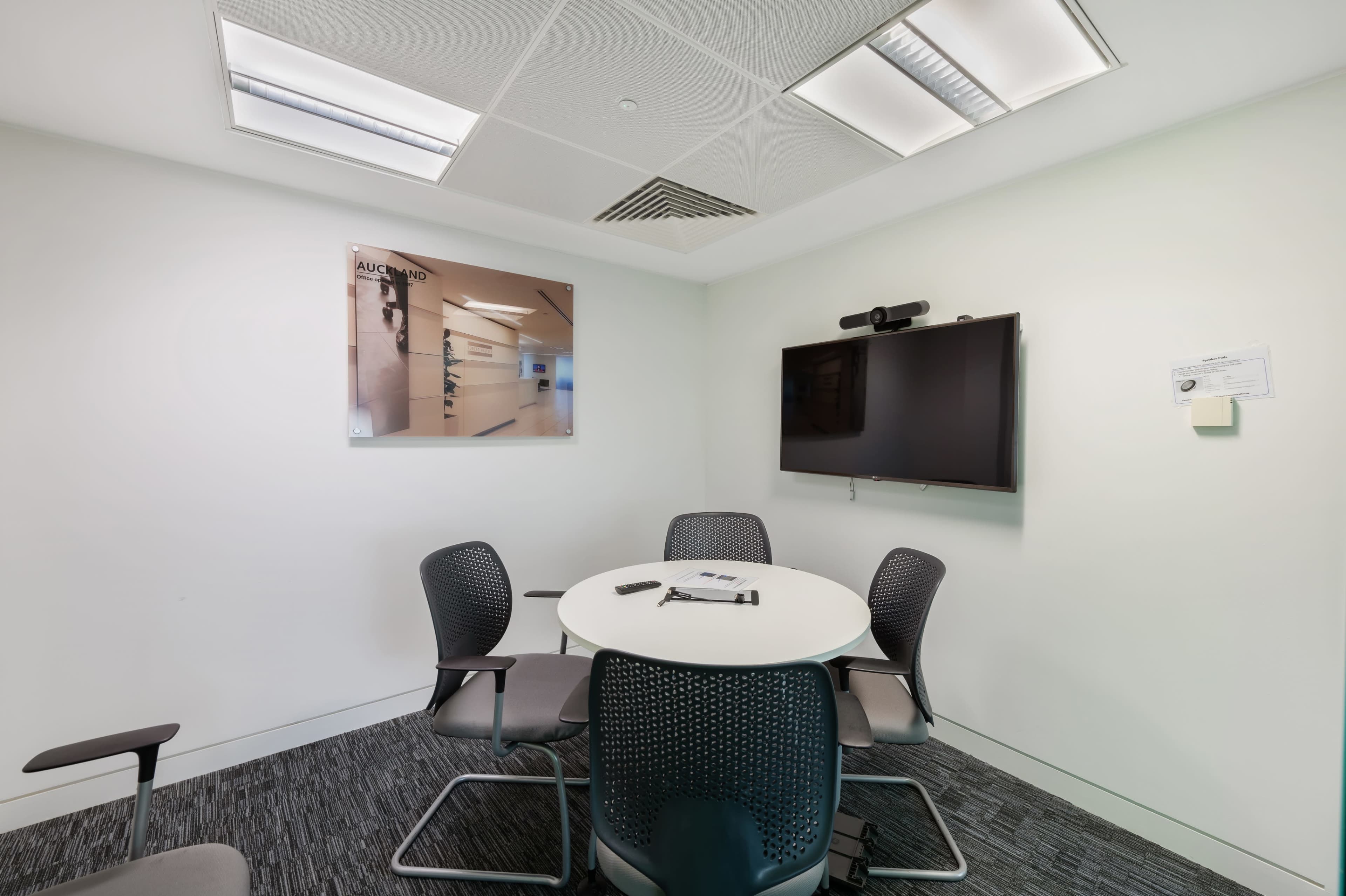 A small conference room features a round table with four chairs, a large wall-mounted screen, and a framed picture on the wall.