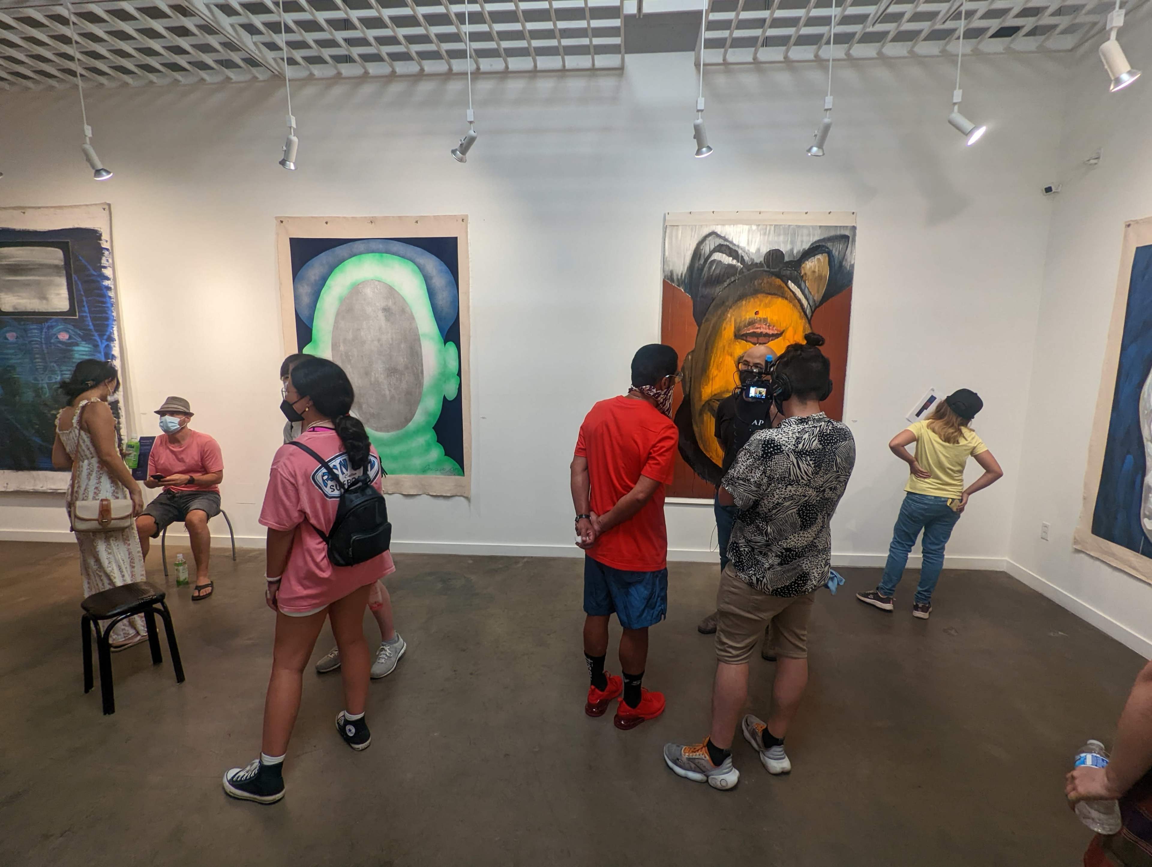 An art gallery featuring several people viewing large paintings on the walls, while others are sitting or engaged in conversation.