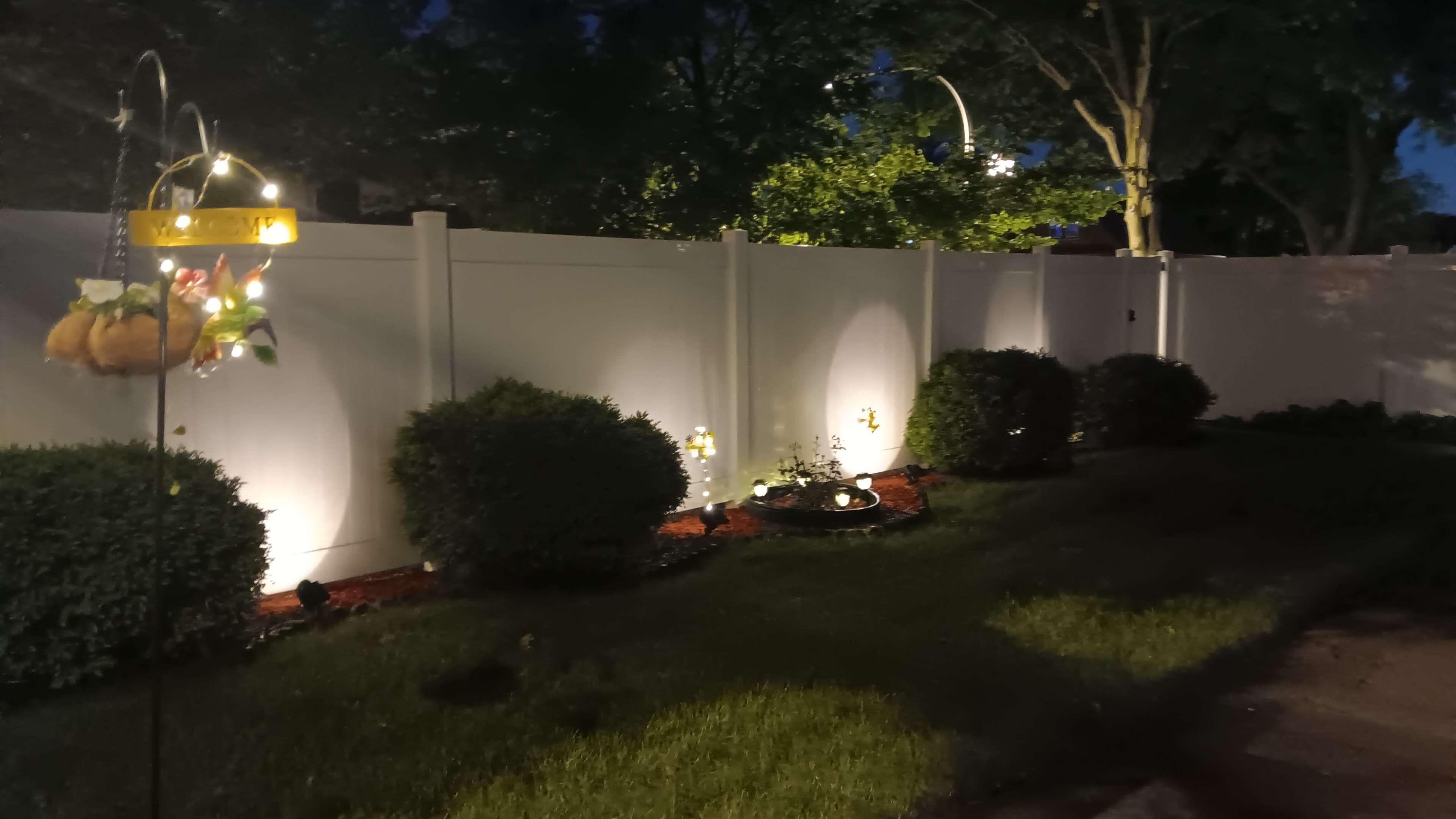 A well-lit garden area at night, featuring a white fence, neatly trimmed bushes, and decorative light fixtures.