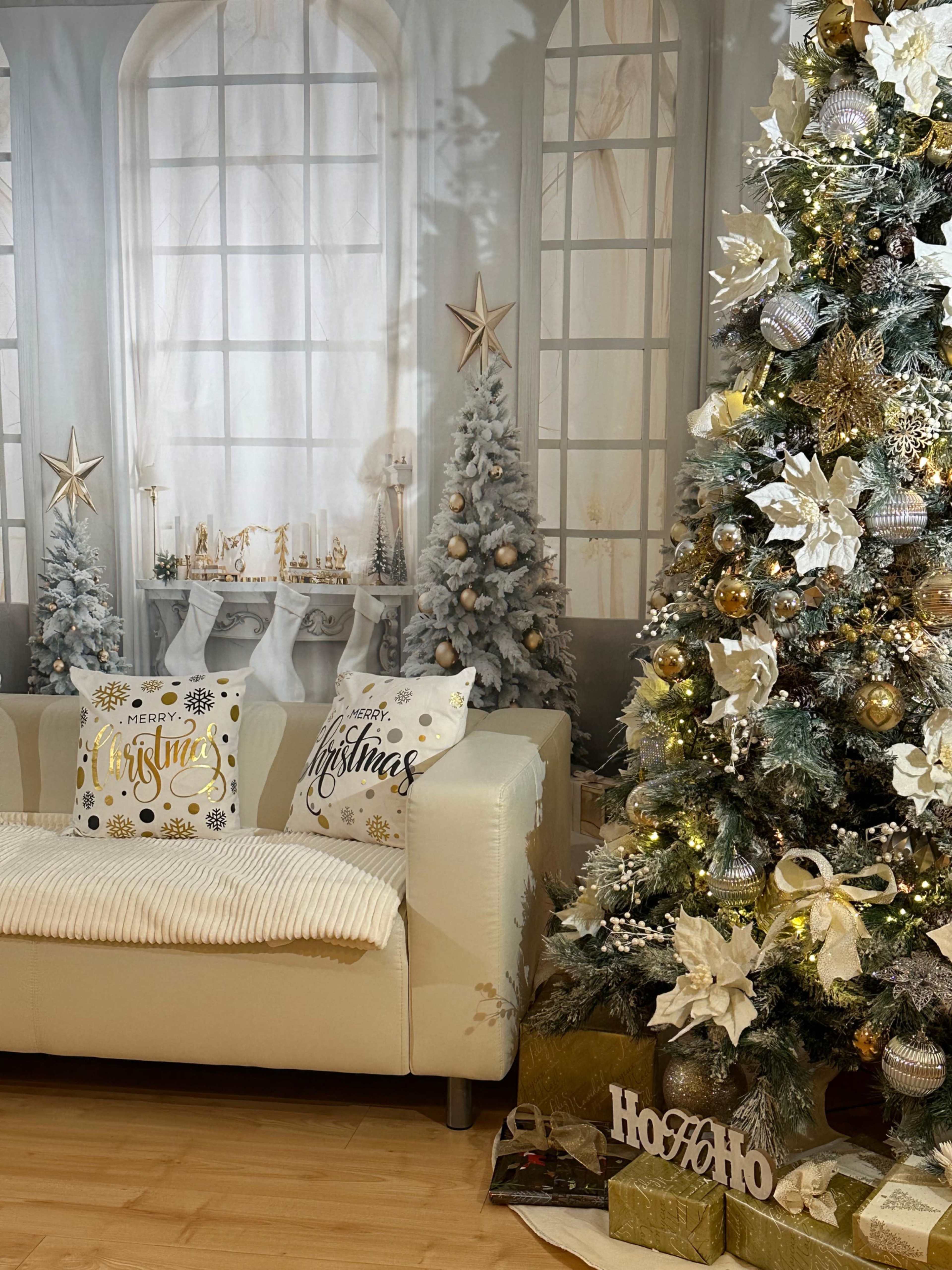The image shows a cozy living room decorated for Christmas, featuring a white sofa with festive pillows, a large Christmas tree adorned with ornaments, and two smaller trees in the background.
