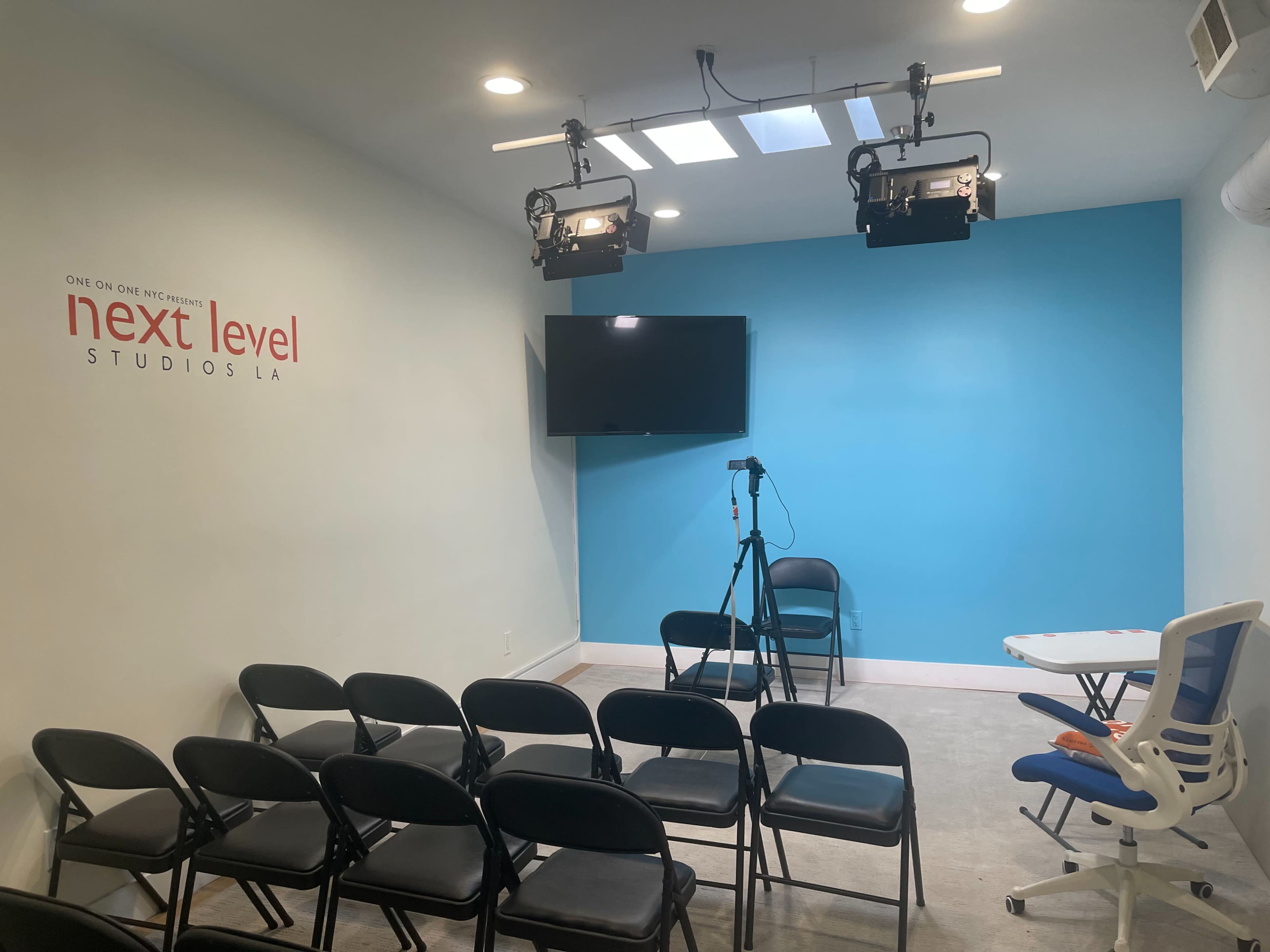 The image shows a small studio with a blue accent wall, a television, a camera on a tripod, and a row of black folding chairs facing the setup.