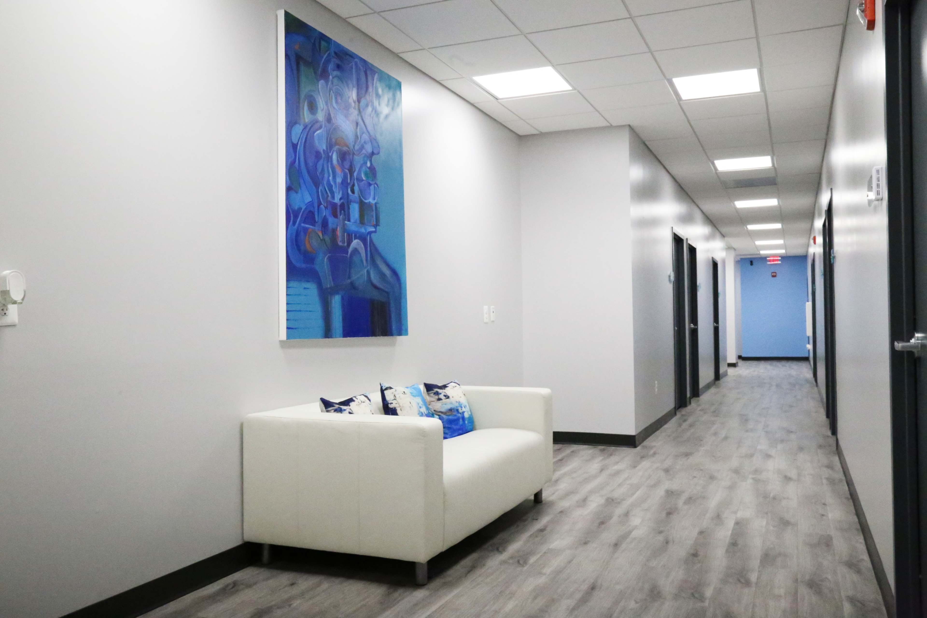 A white sofa with decorative pillows sits against a wall in a long, well-lit hallway featuring several doors and a large piece of abstract art.