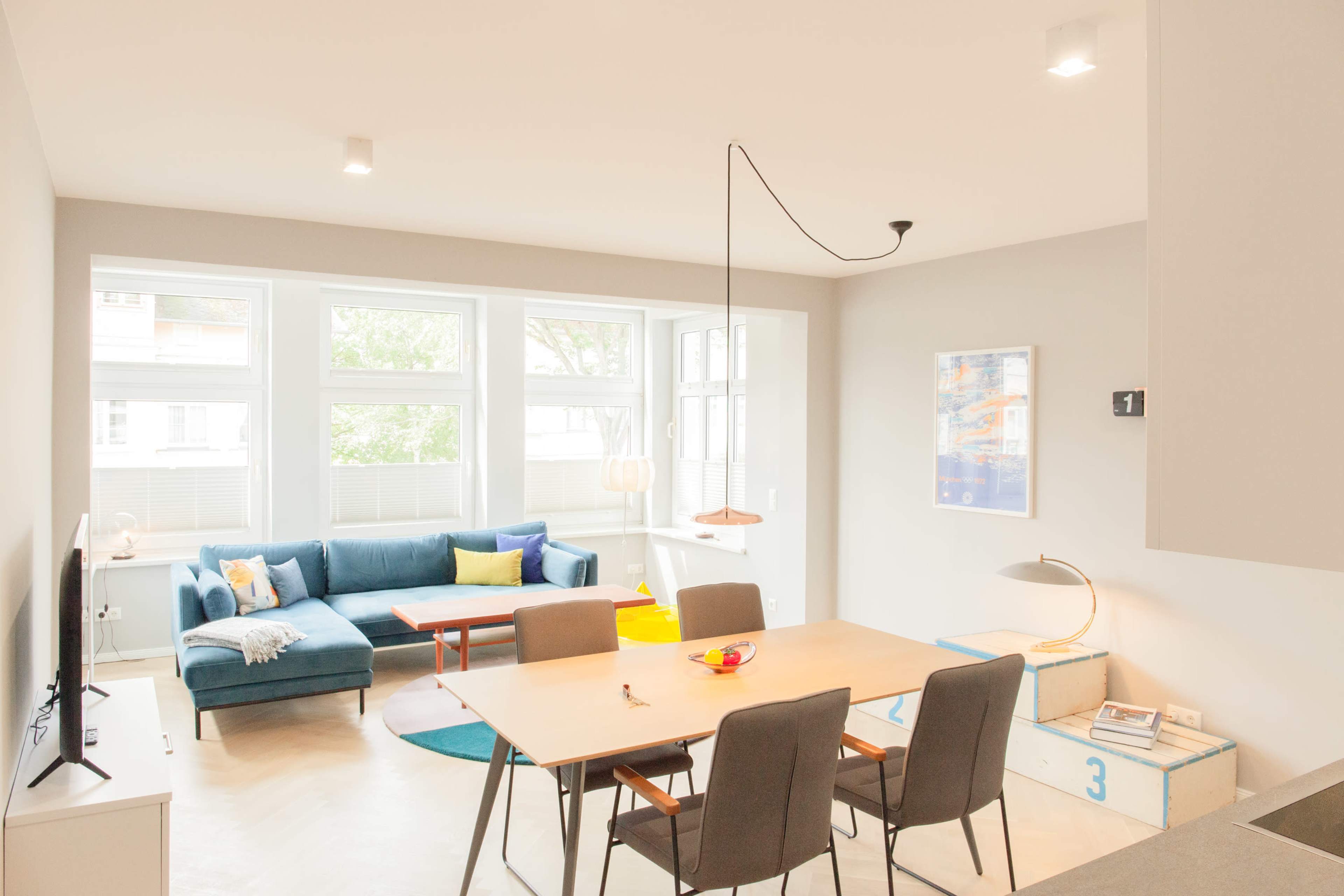 A modern living and dining area with a blue sofa, a wooden dining table, and large windows that let in natural light.