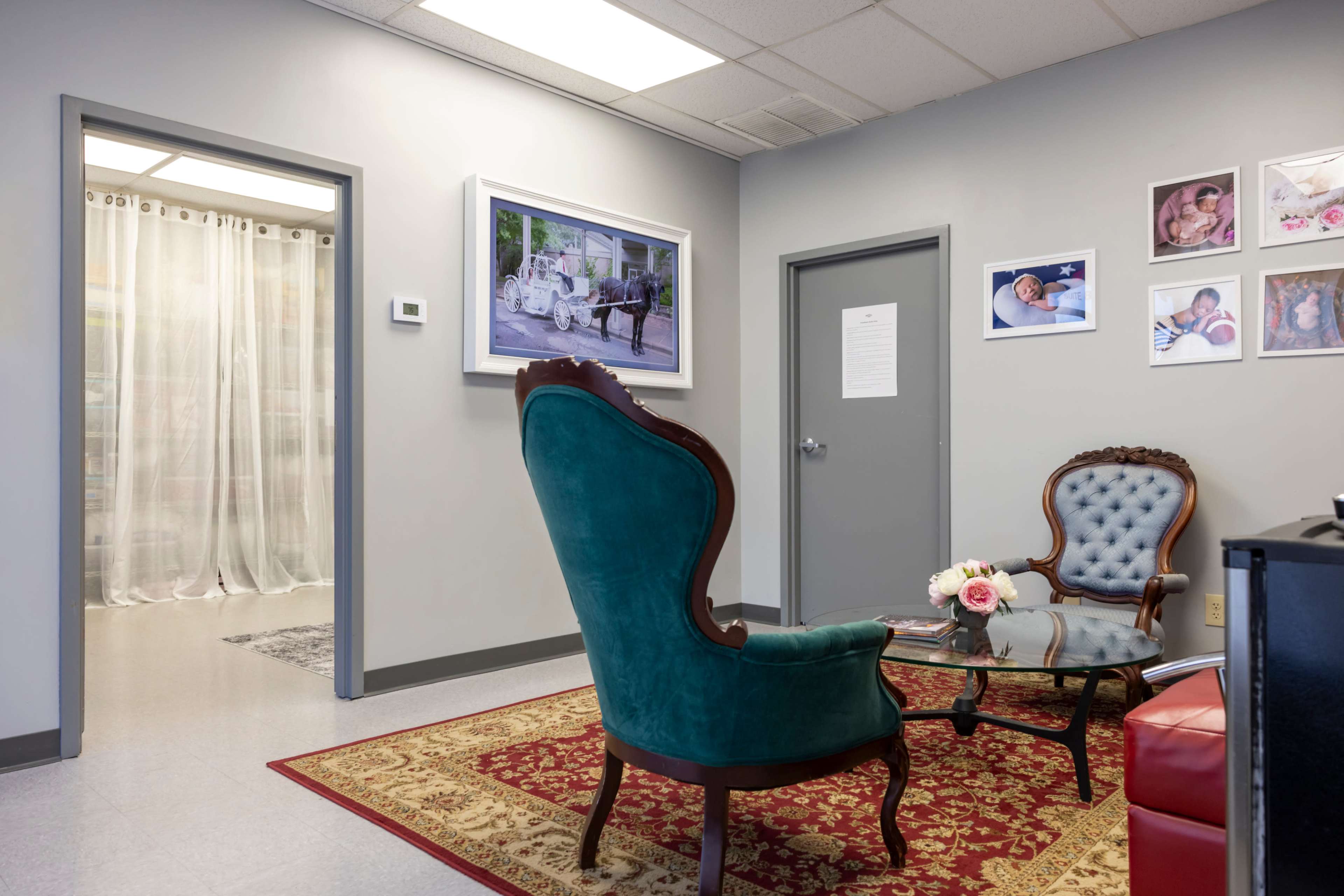 The image shows a waiting area with two upholstered chairs, a small table with flowers, and framed photographs on the walls.