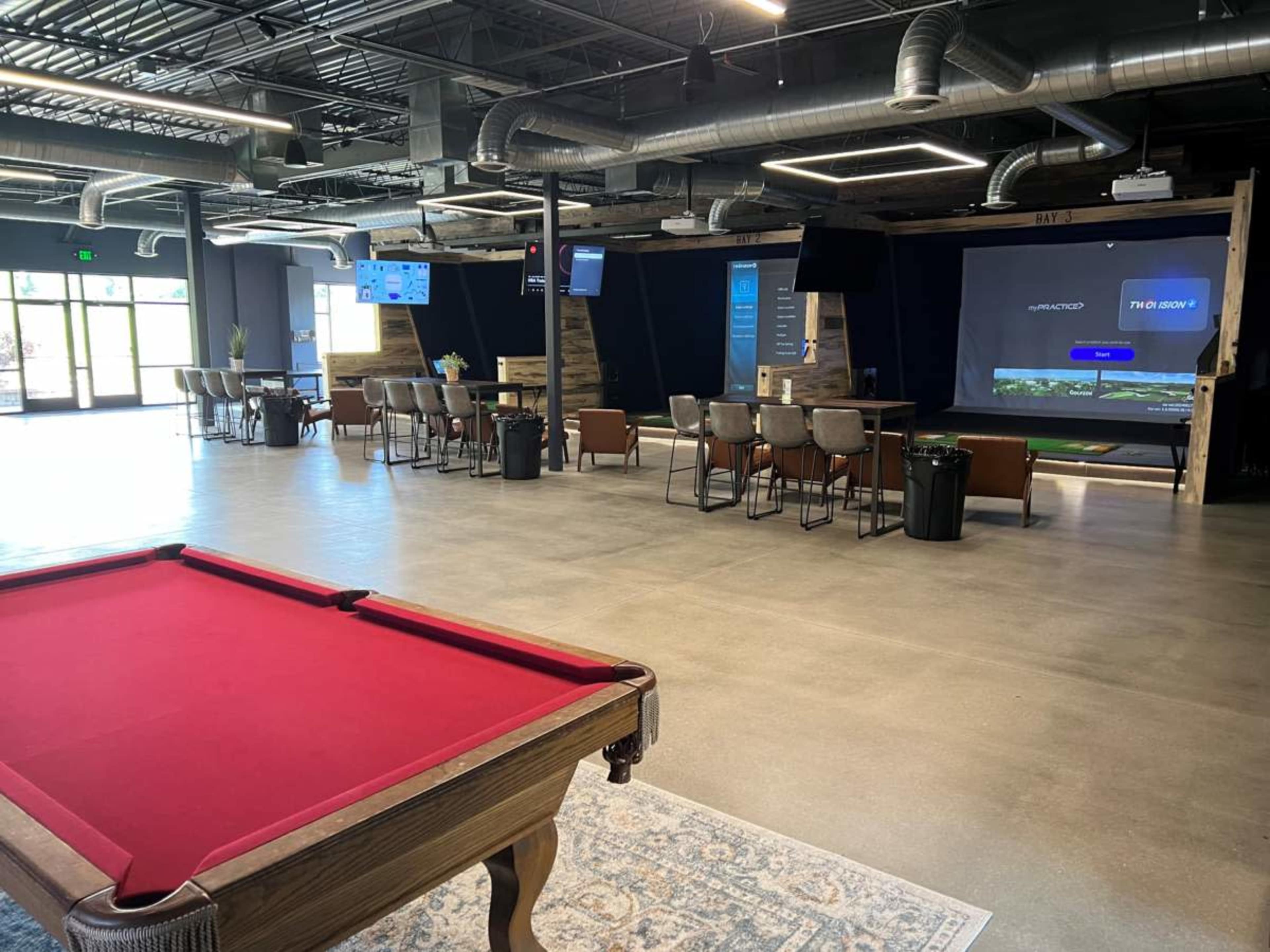 The image shows an open recreational space featuring a pool table in the foreground, with seating areas and screens visible in the background.