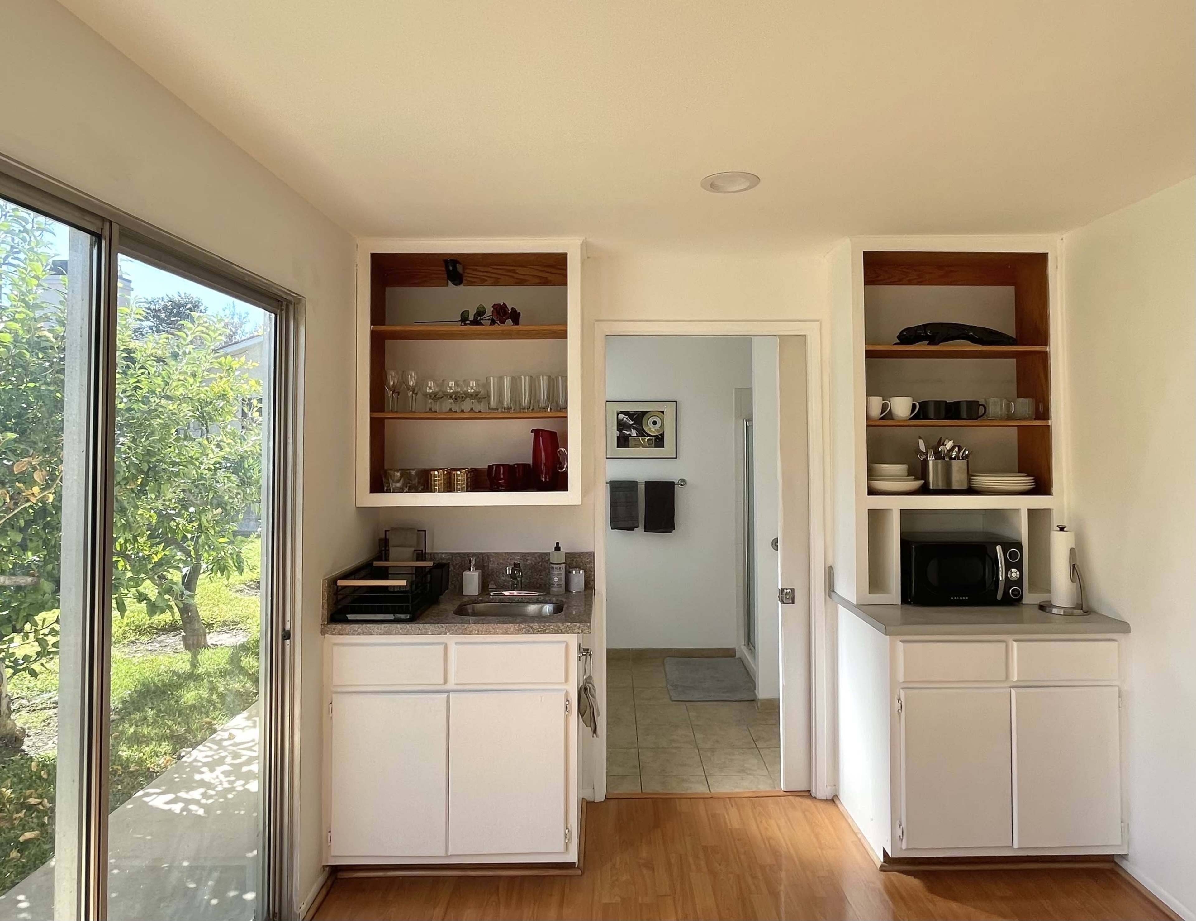 A kitchen space with open shelves, a sink, microwave, and large windows overlooking a garden.