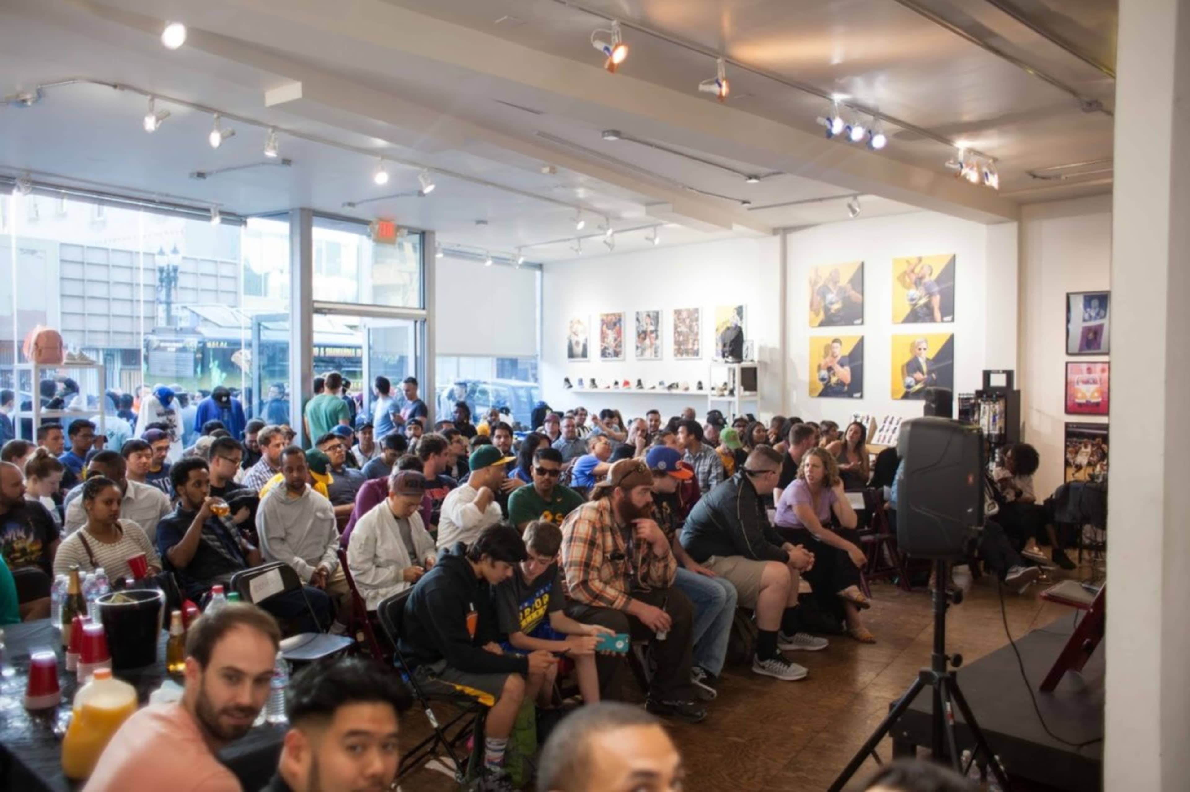 A large crowd fills a modern gallery space, seated and facing a stage at the front.