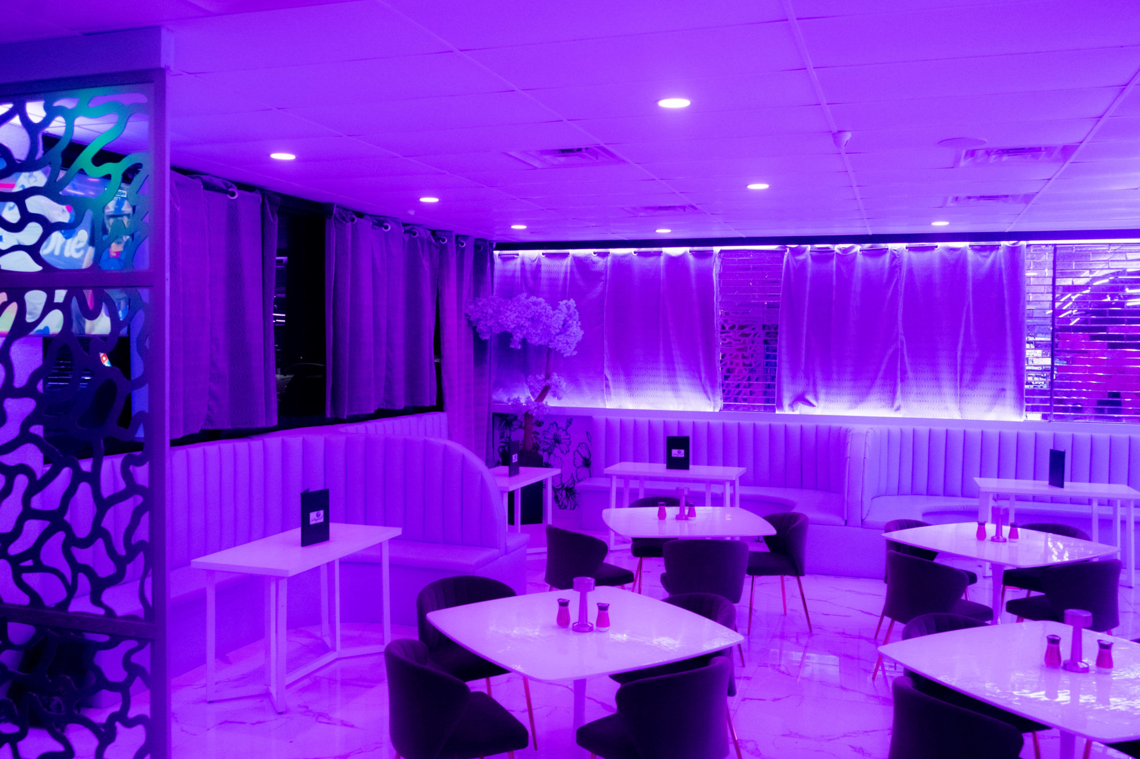 The image shows a modern restaurant interior with purple lighting, featuring several white tables and dark chairs arranged around the space.