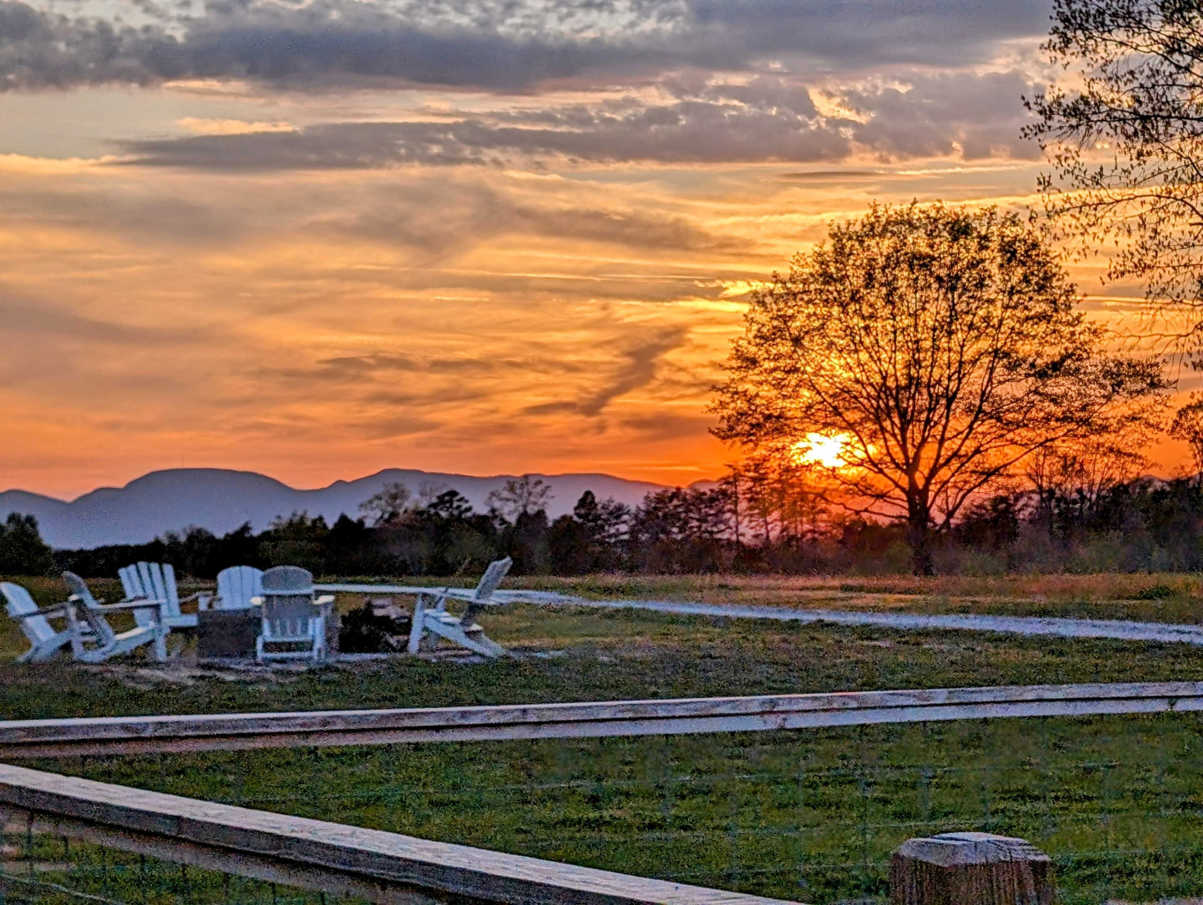 A sunset over a mountainous landscape with Adirondack chairs arranged around a fire pit in the foreground.