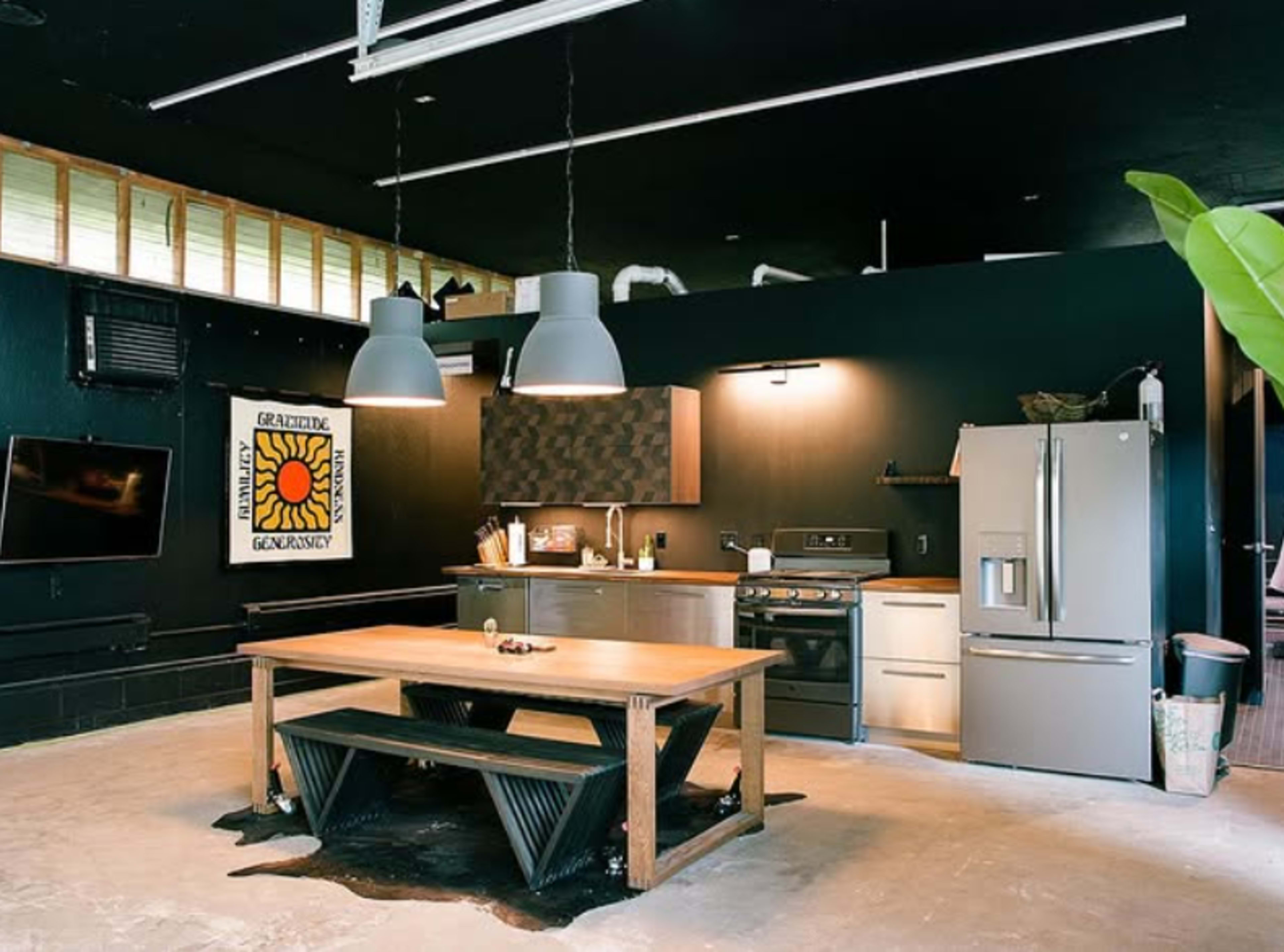 The image shows a modern kitchen with a large wooden table, stainless steel appliances, and black walls with a decorative wall hanging.