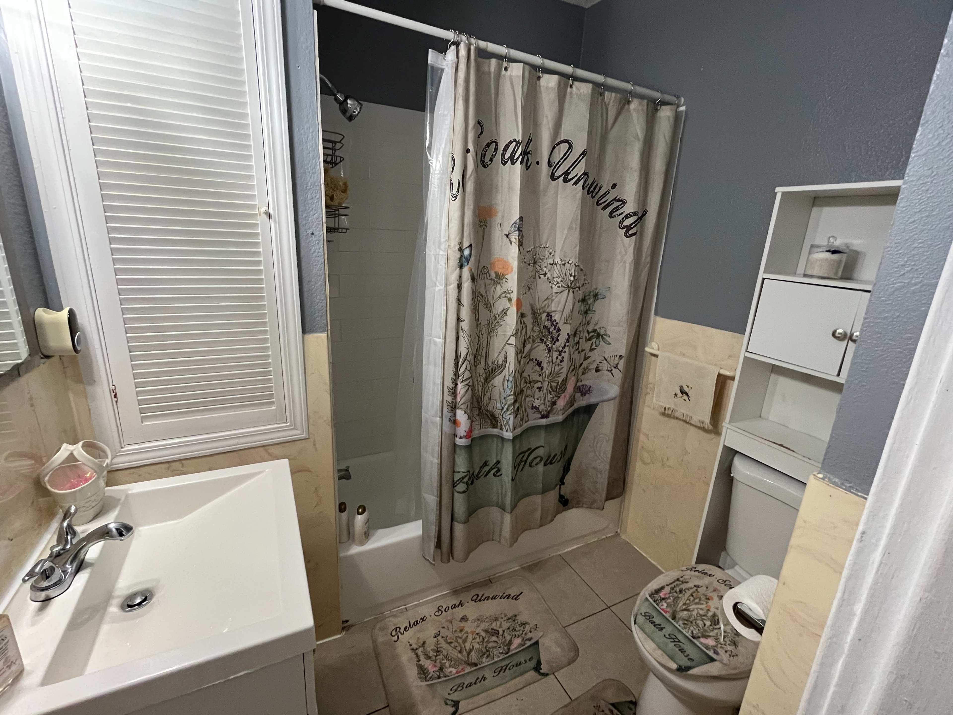 A bathroom features a shower with a floral curtain, a sink with a cabinet, and a toilet, all surrounded by tiled walls.