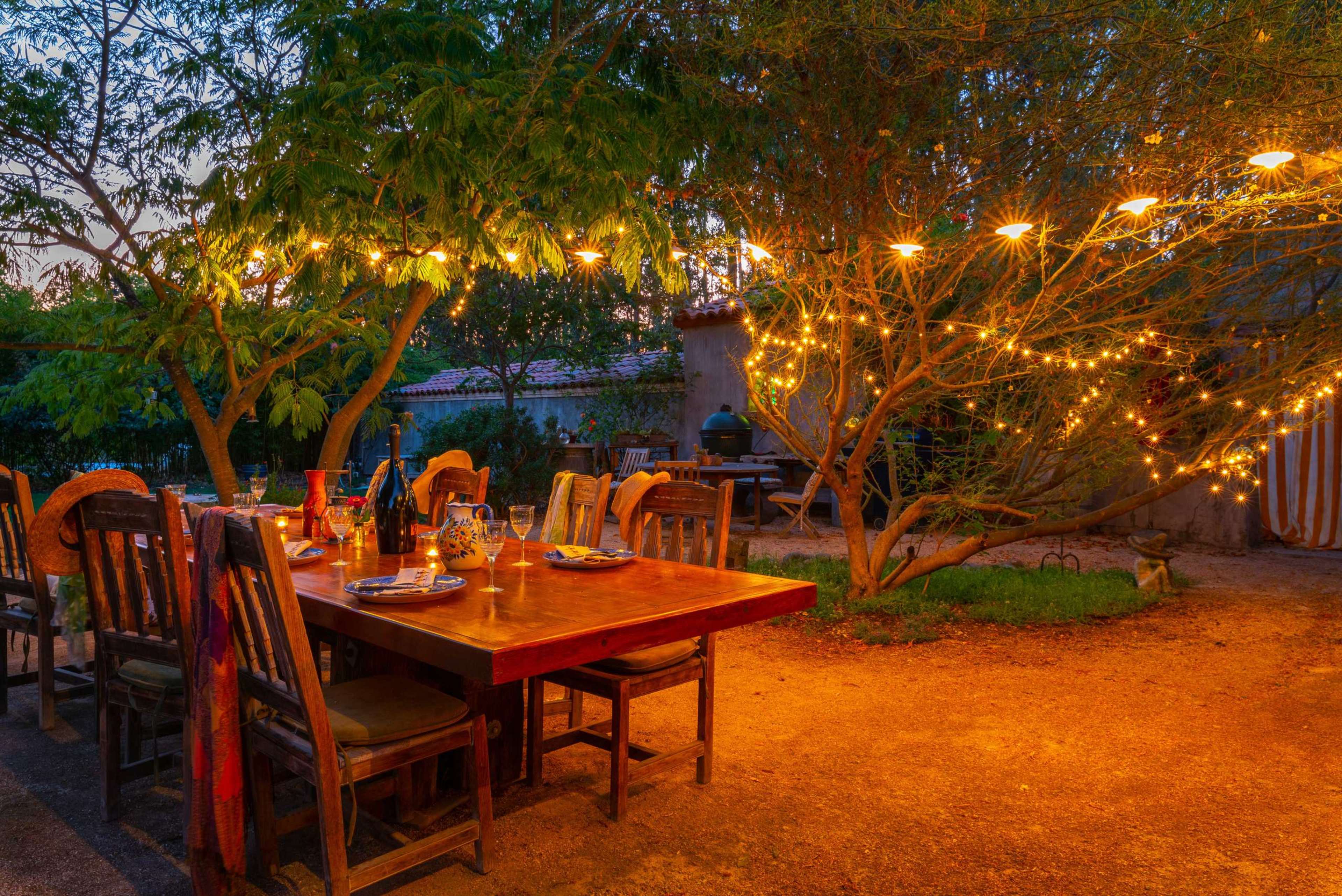 A wooden dining table is set outdoors under string lights, surrounded by chairs and a tree, in a warm, twilight setting.