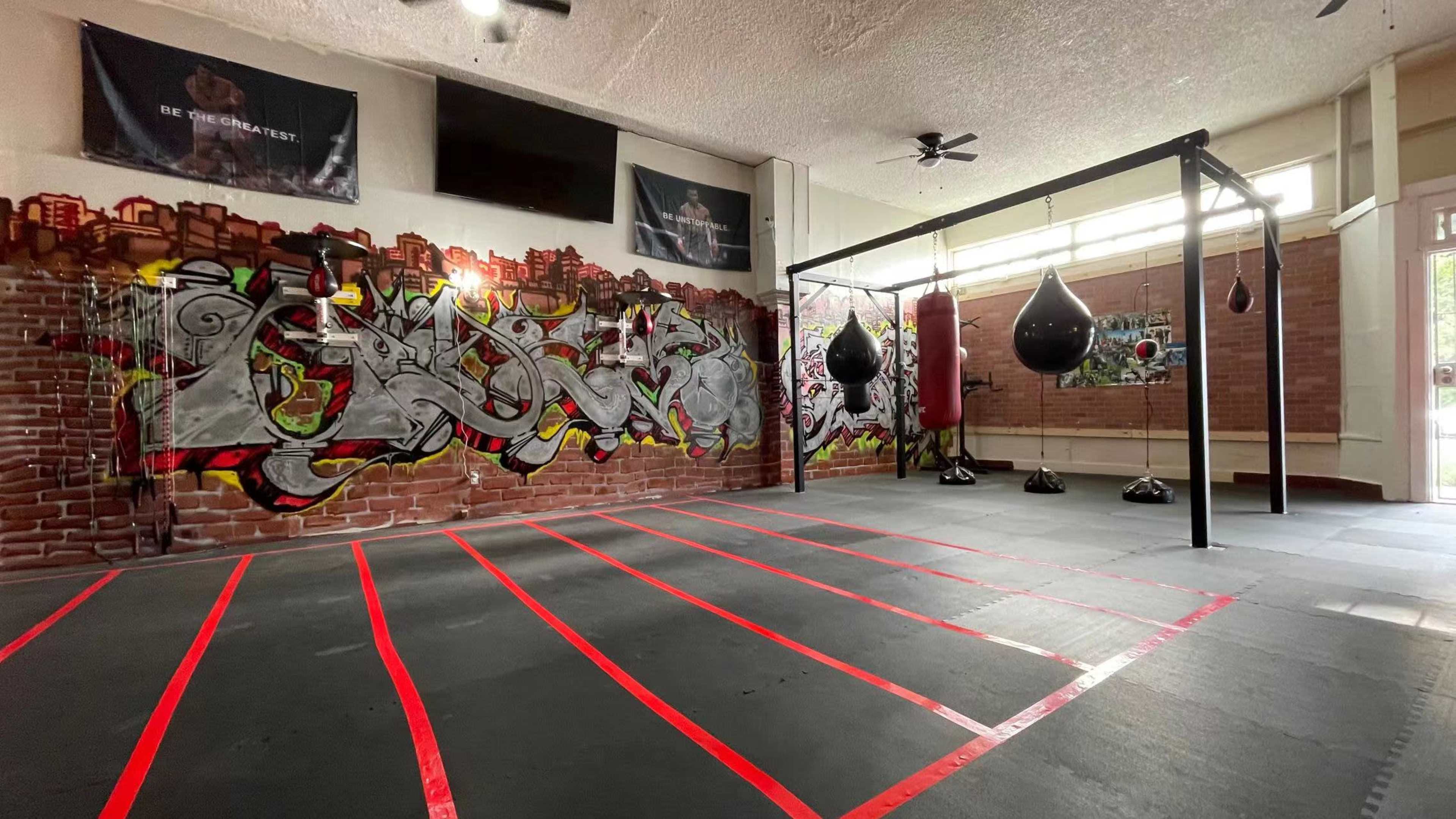 An indoor gym with a graffiti-covered wall, a boxing ring, and several hanging punching bags.