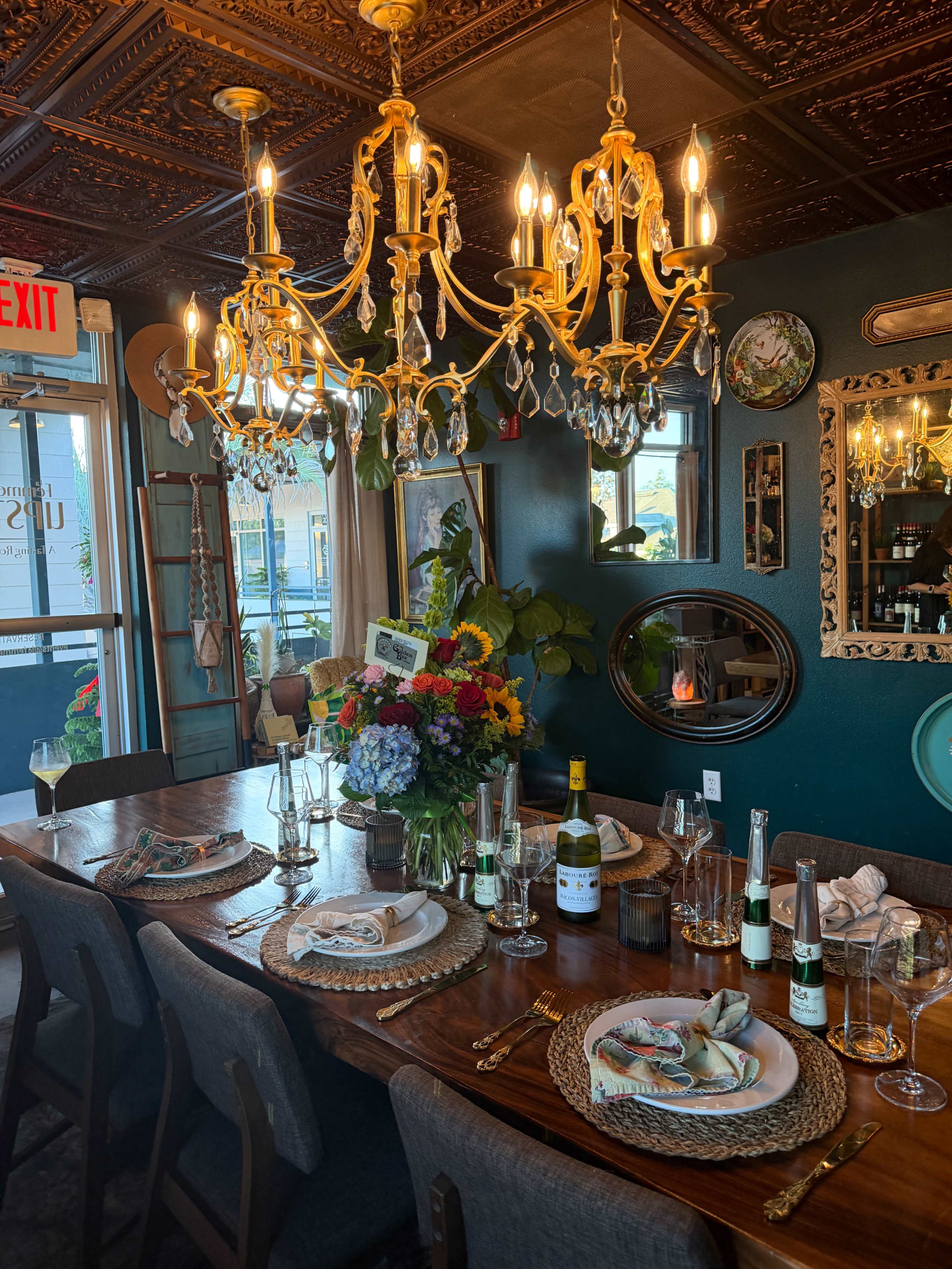A dining room table is set with elegant plates and glassware, adorned with a vibrant floral arrangement, beneath a decorative chandelier.