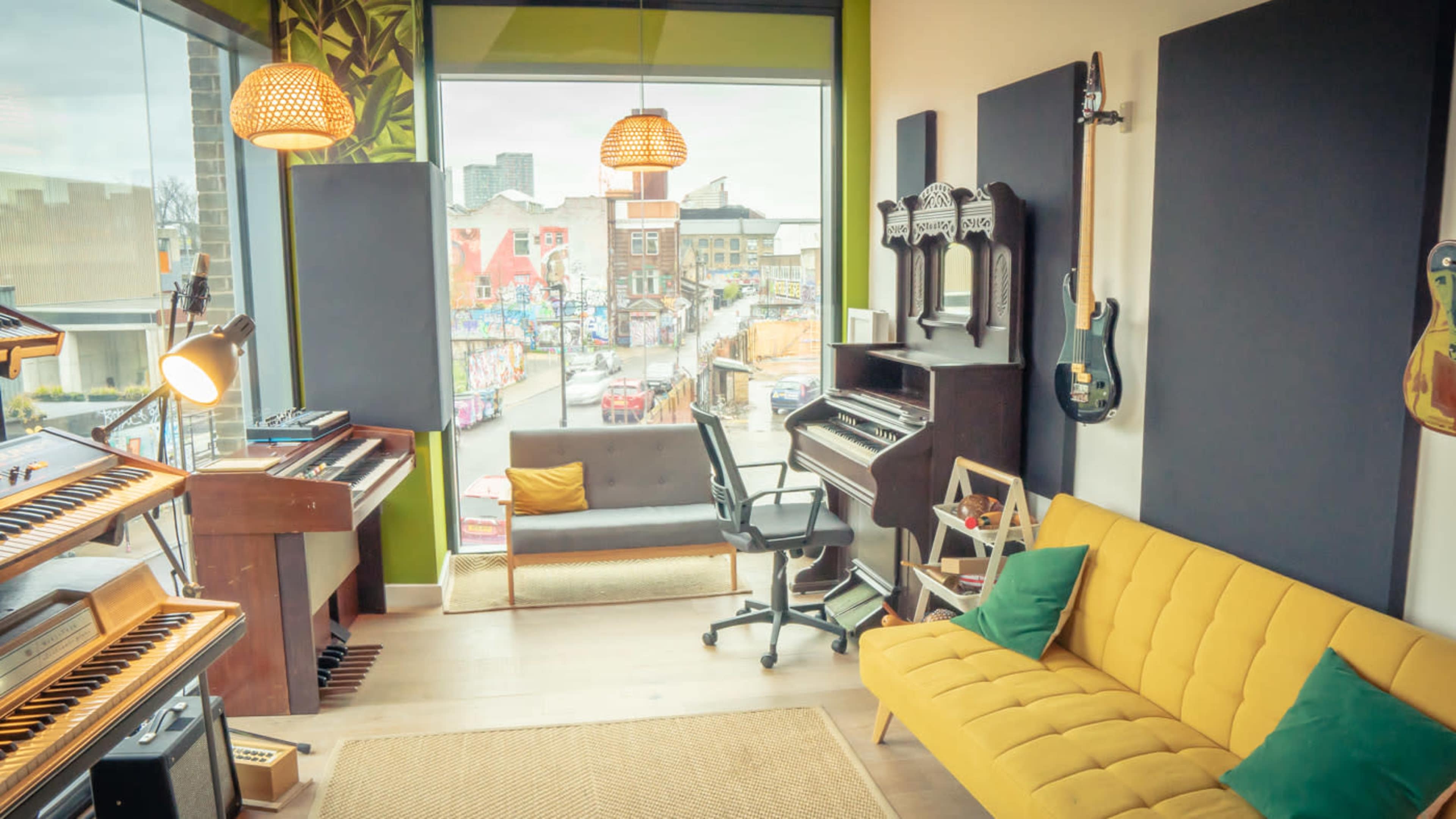 The image shows a modern music studio with a yellow sofa, a desk with a chair, and various musical instruments visible through large windows overlooking a street with graffiti art.