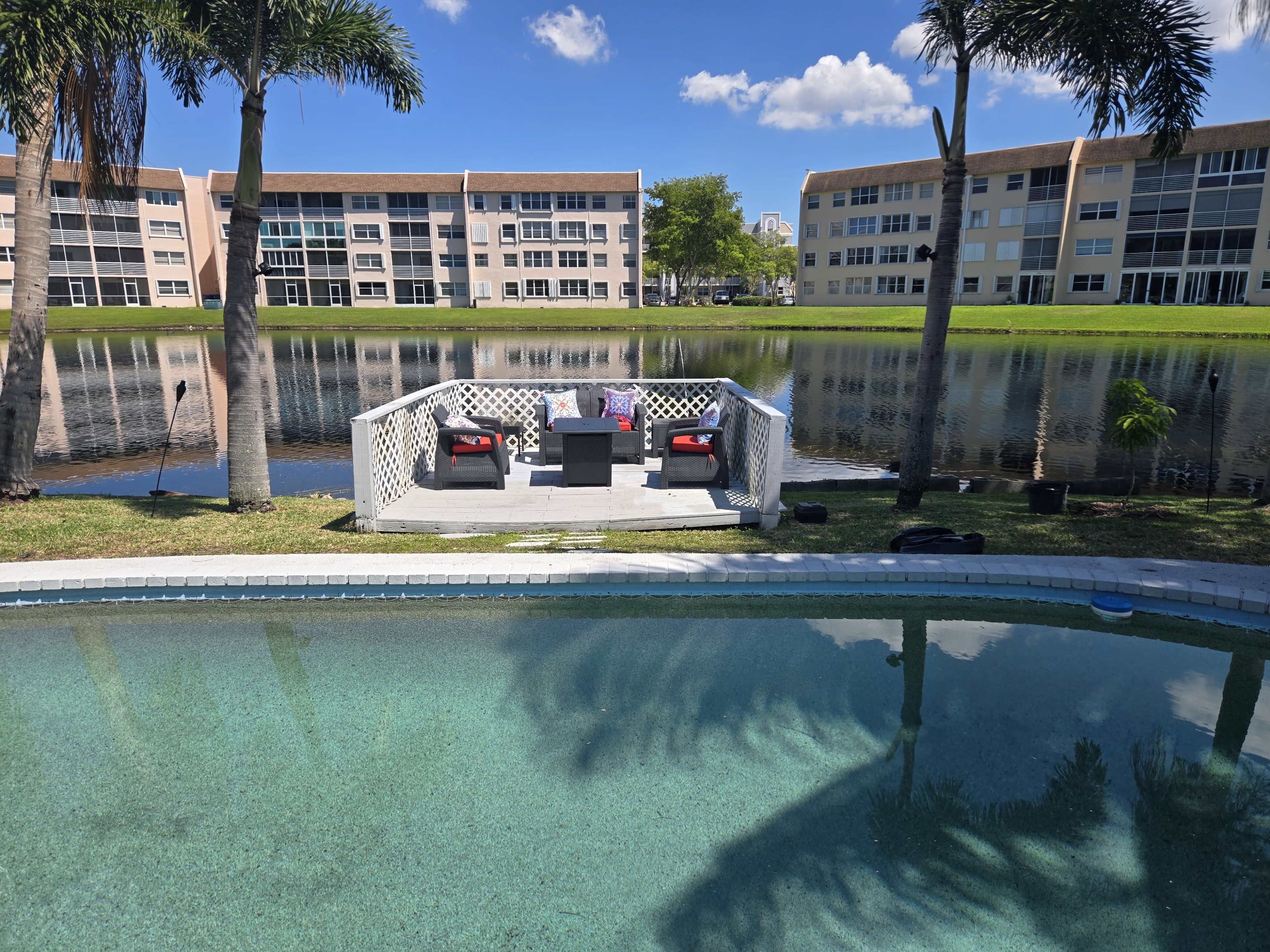 A dock with seating overlooks a lake, bordered by a swimming pool and surrounded by palm trees and residential buildings.