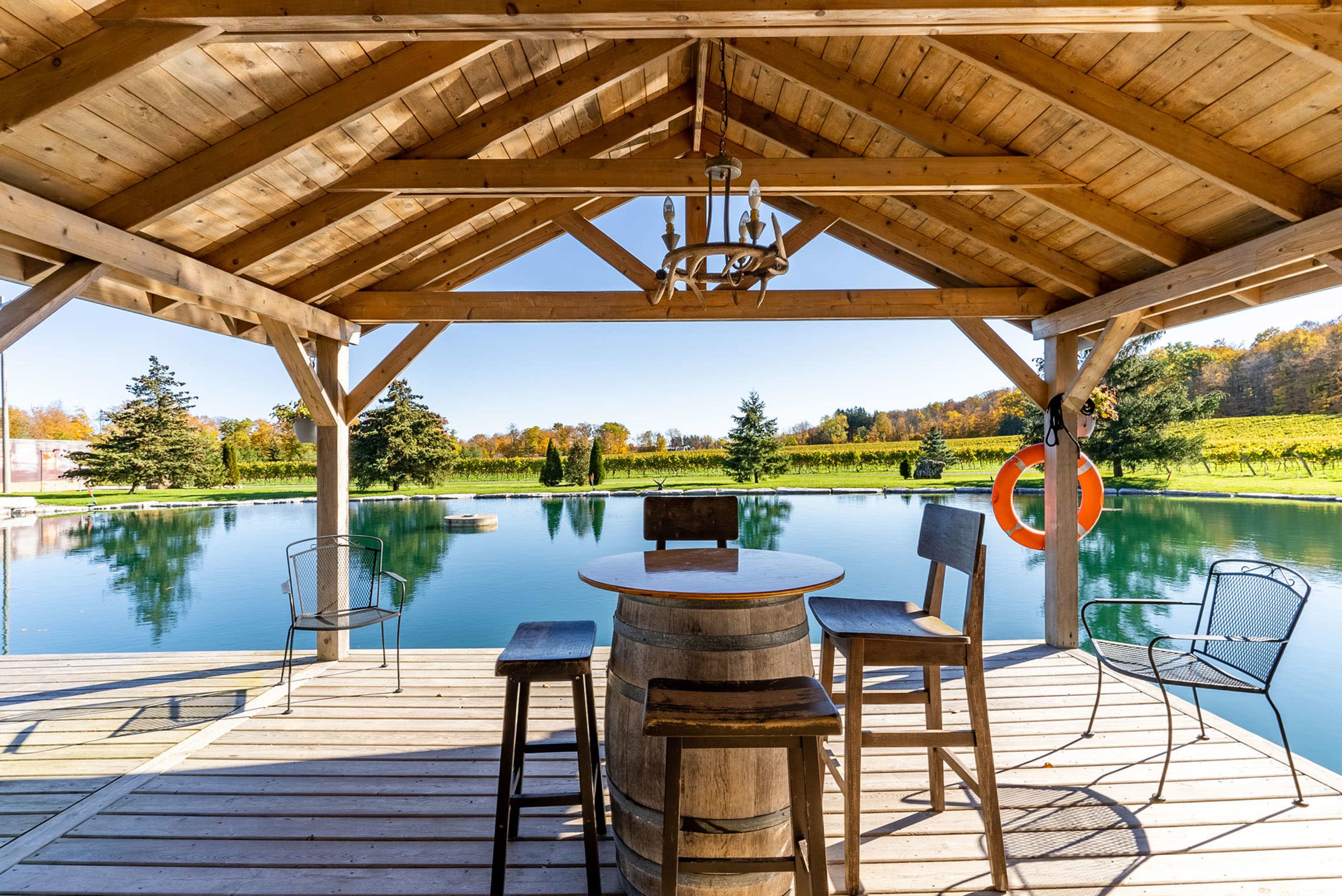 A wooden pavilion with a circular table and chairs overlooks a calm, reflective body of water surrounded by greenery.