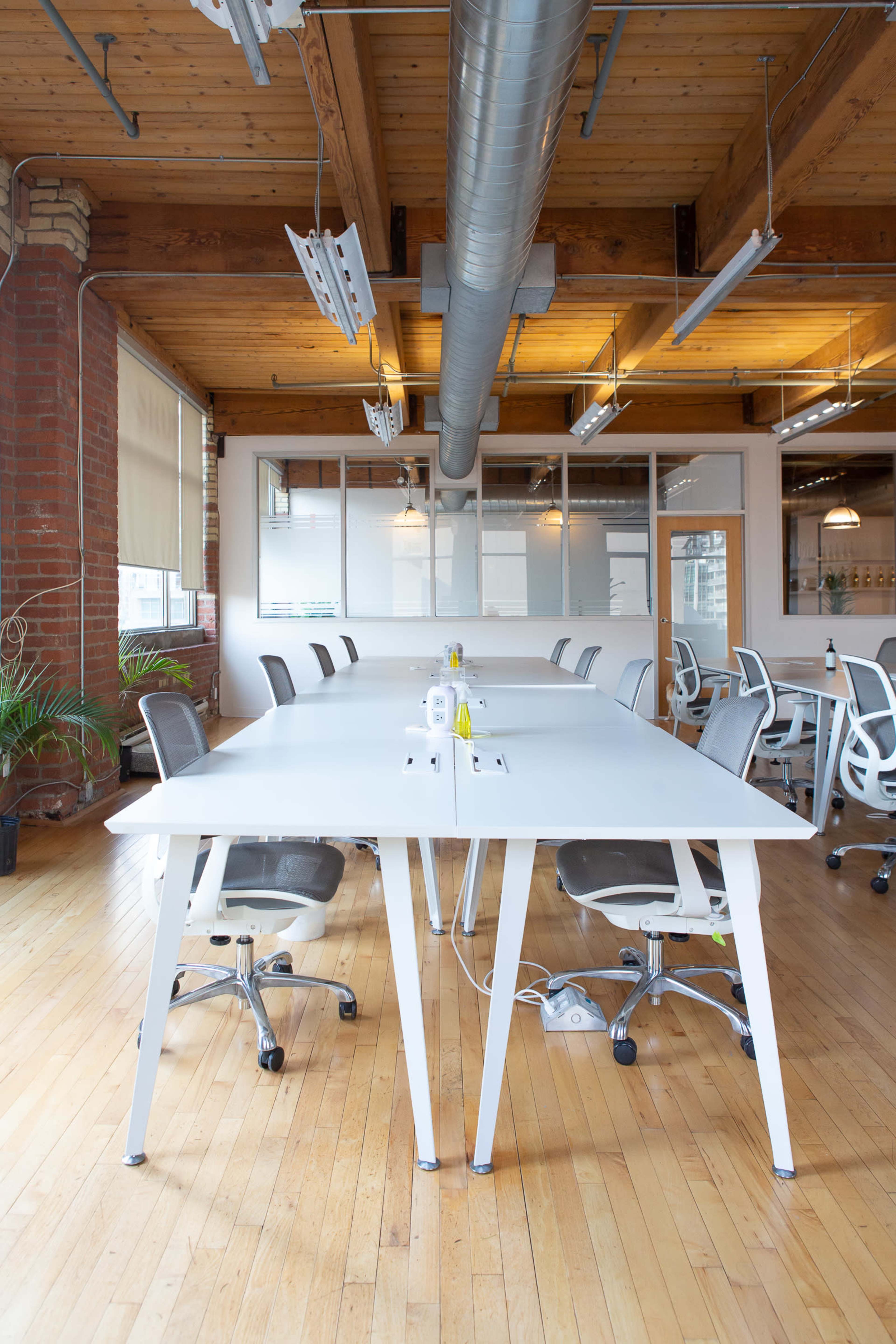 A modern office space features a large white table surrounded by ergonomic chairs, with wooden floors and exposed brick walls.