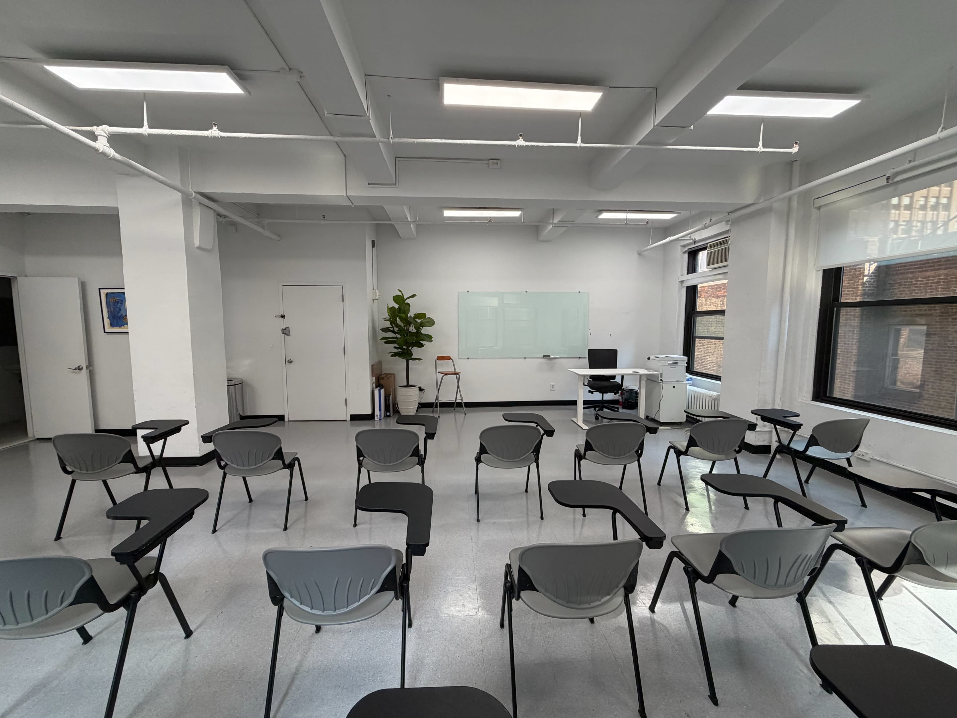 The image shows a classroom setup with rows of empty chairs facing a whiteboard and a desk on one side of the room.