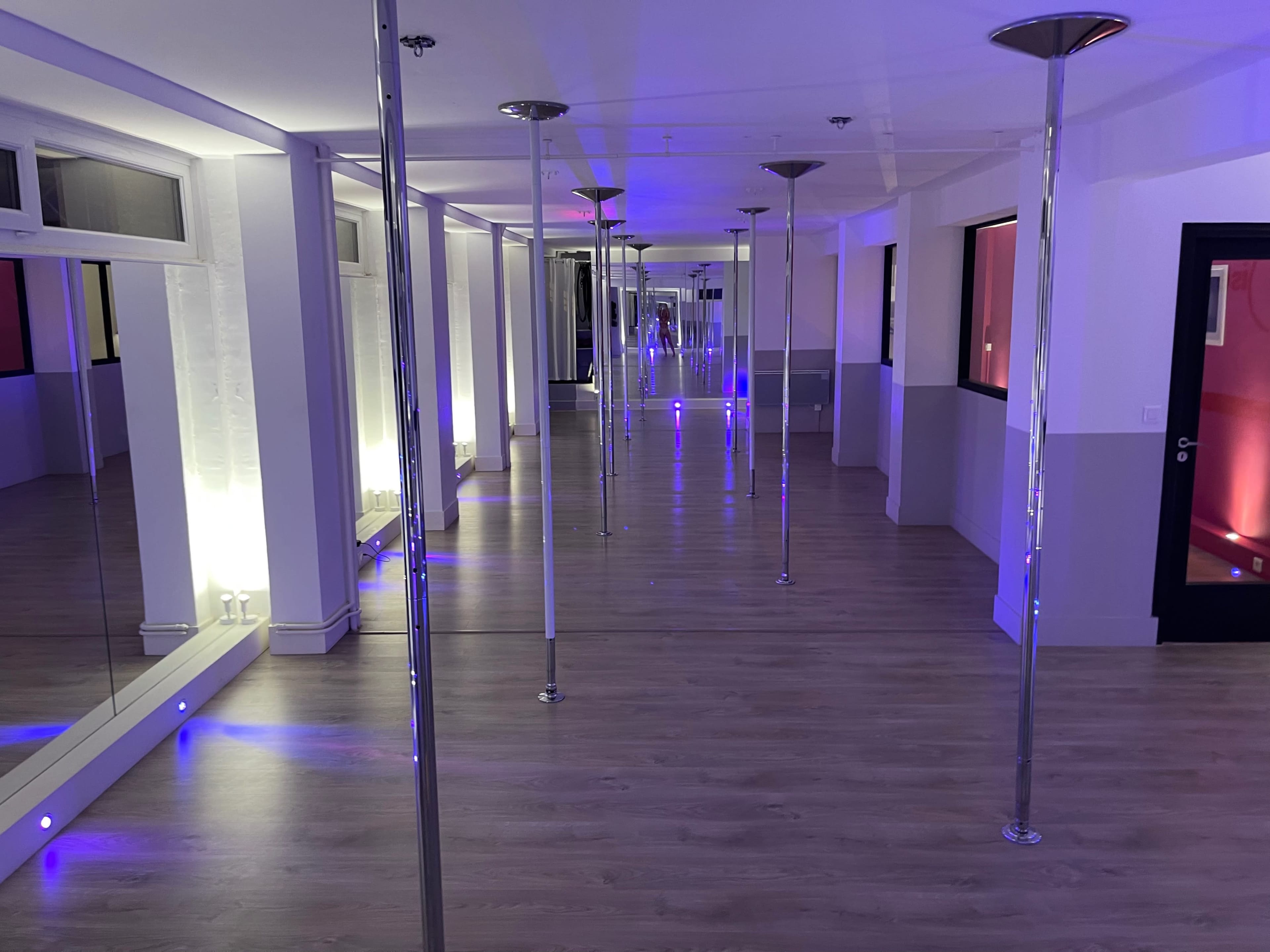 A spacious dance studio with multiple vertical poles, mirrored walls, and blue lighting creating an ambient atmosphere.