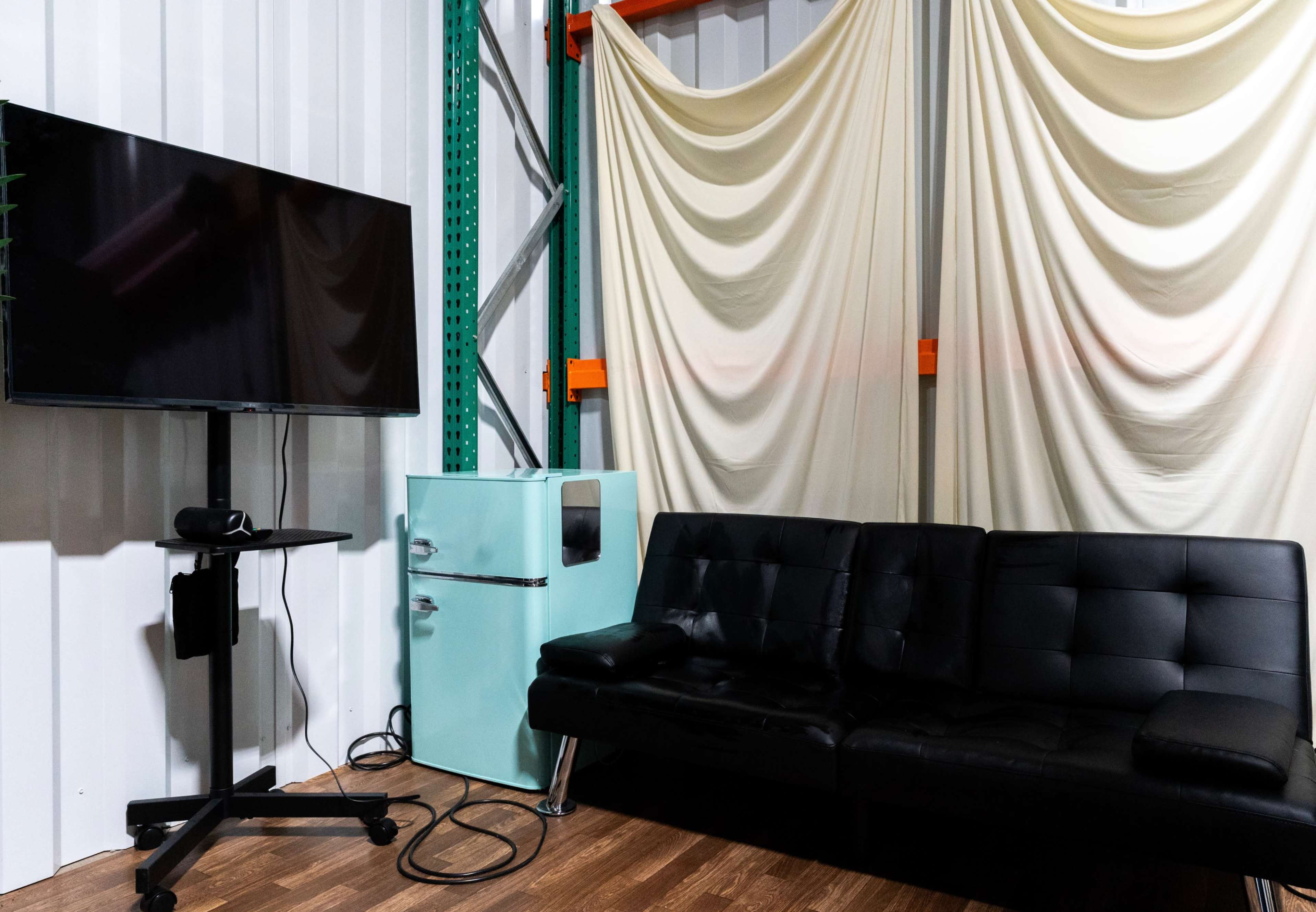 The image shows a small lounge area featuring a black couch, a TV on a stand, and a light blue refrigerator against a backdrop of draped fabric.