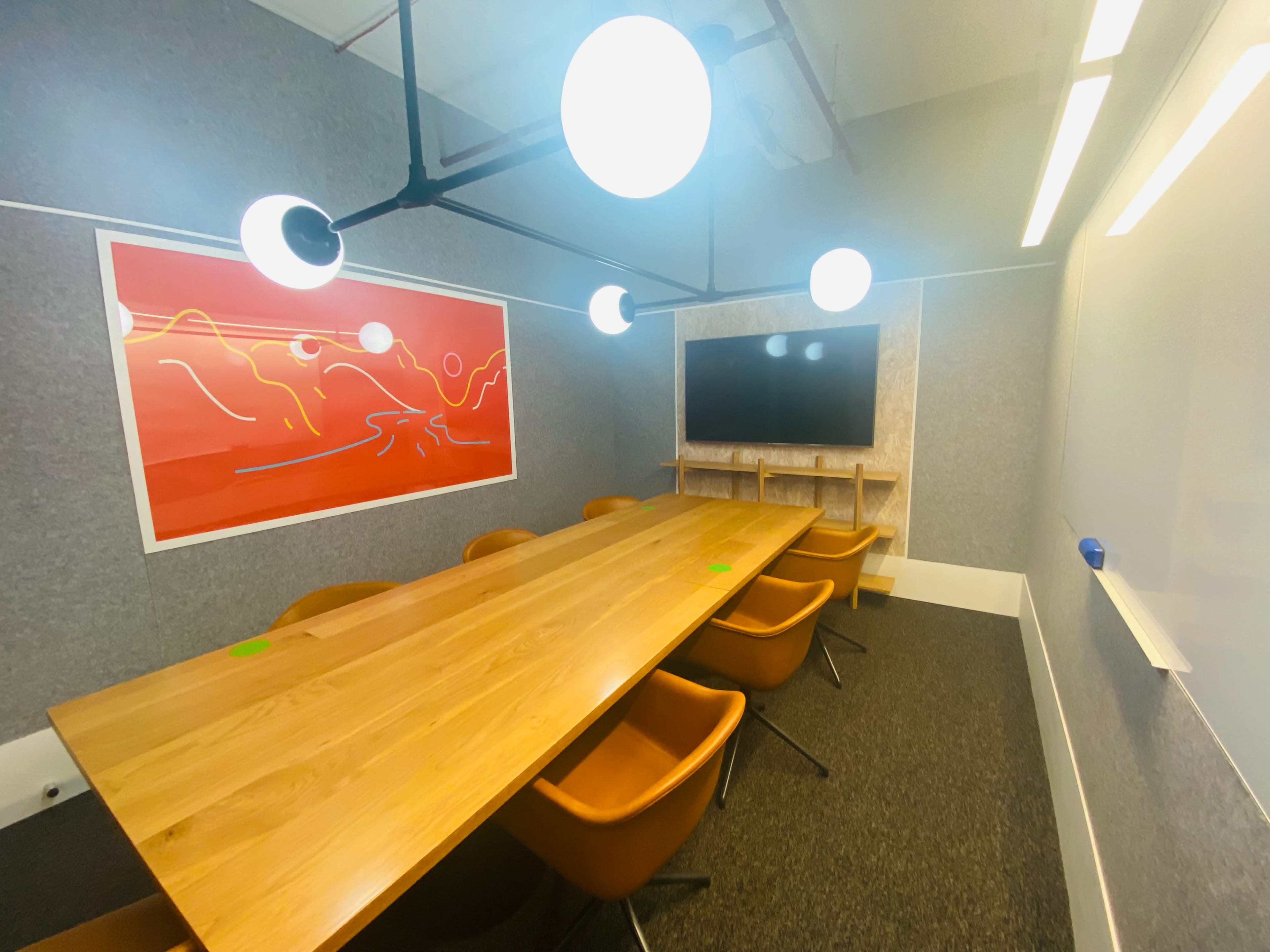 A modern conference room features a long wooden table surrounded by orange chairs, with a large screen mounted on the wall and bright overhead lighting.
