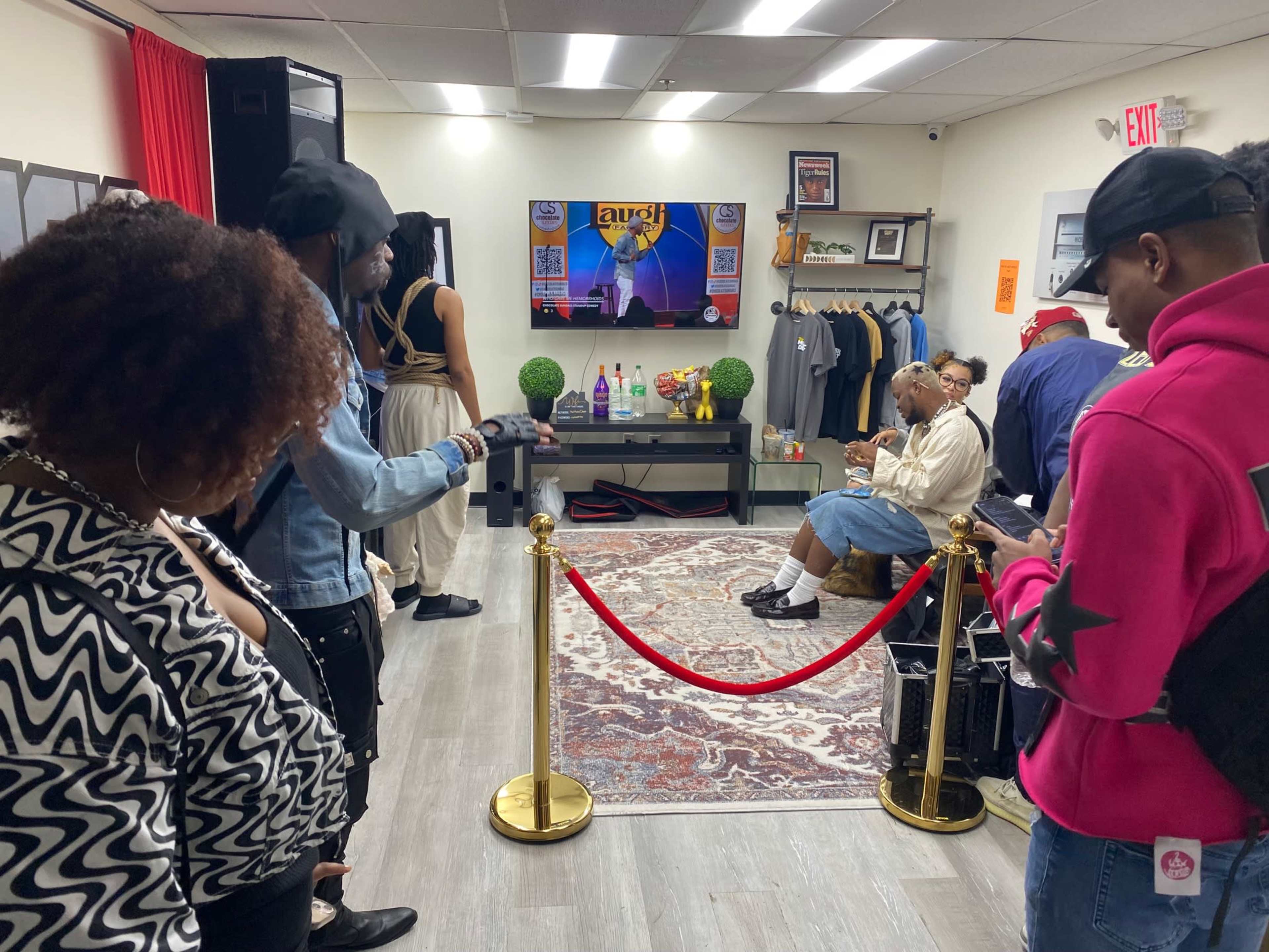 A group of people stands in a retail space with a red carpet leading to a seated individual in front of a television screen and clothing displays.
