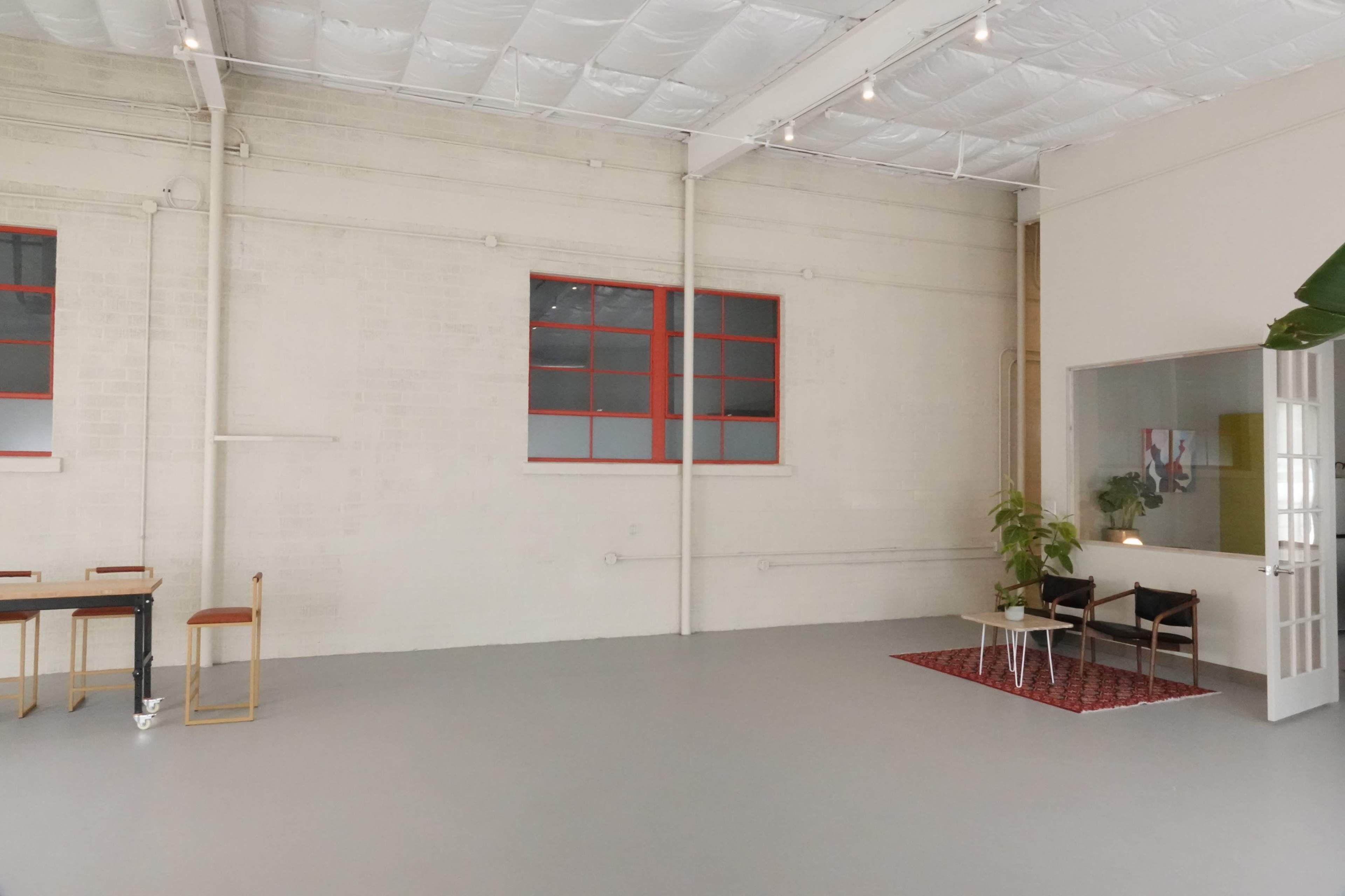 A spacious, empty room with white walls, a gray floor, and red-trimmed windows, featuring a small table with chairs and a rug in one corner.