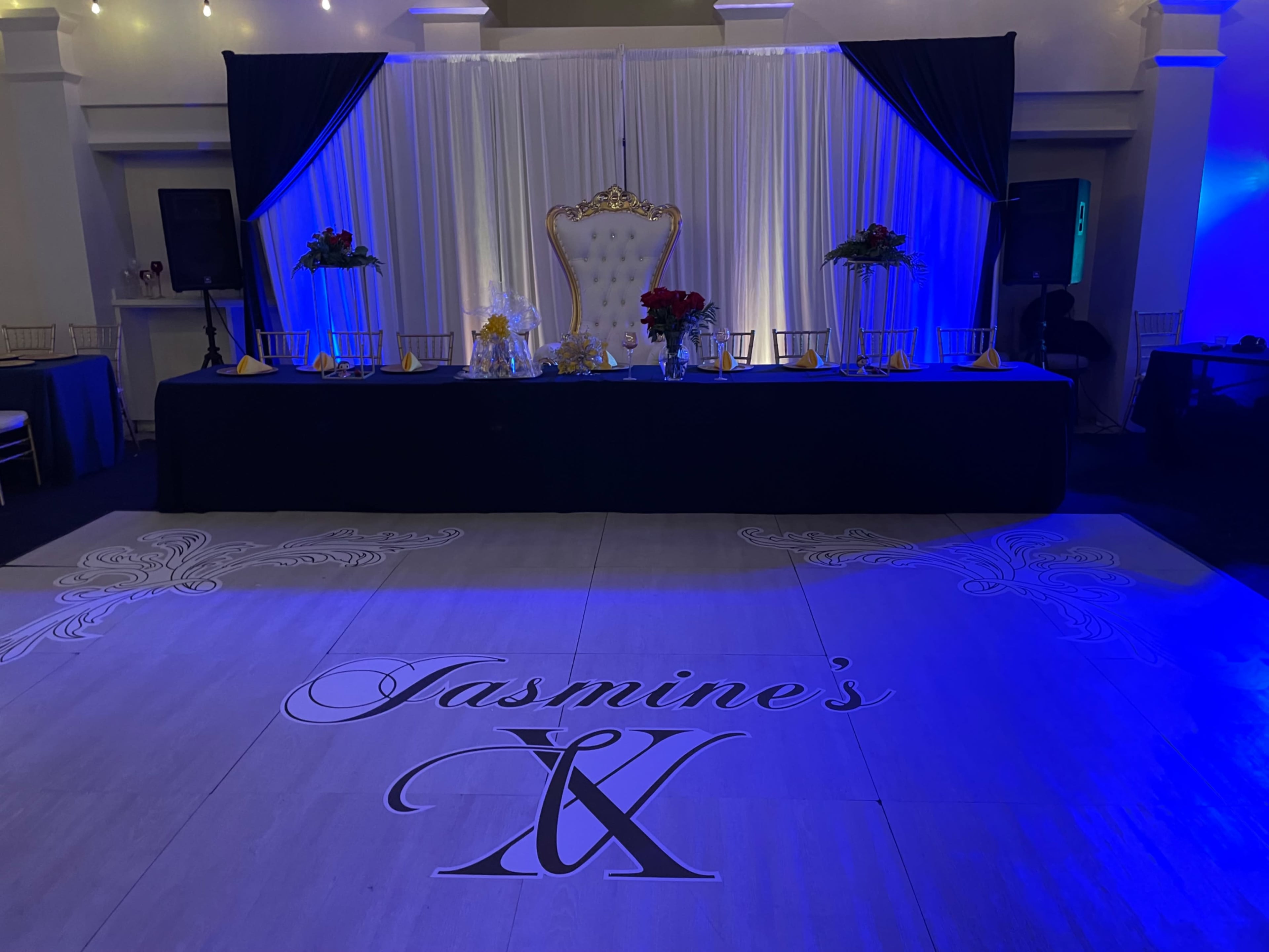 The scene shows a decorated event space featuring a large throne-like chair on a stage, a table set for guests, and a floor design that reads "Jasmine's IX" under soft blue lighting.