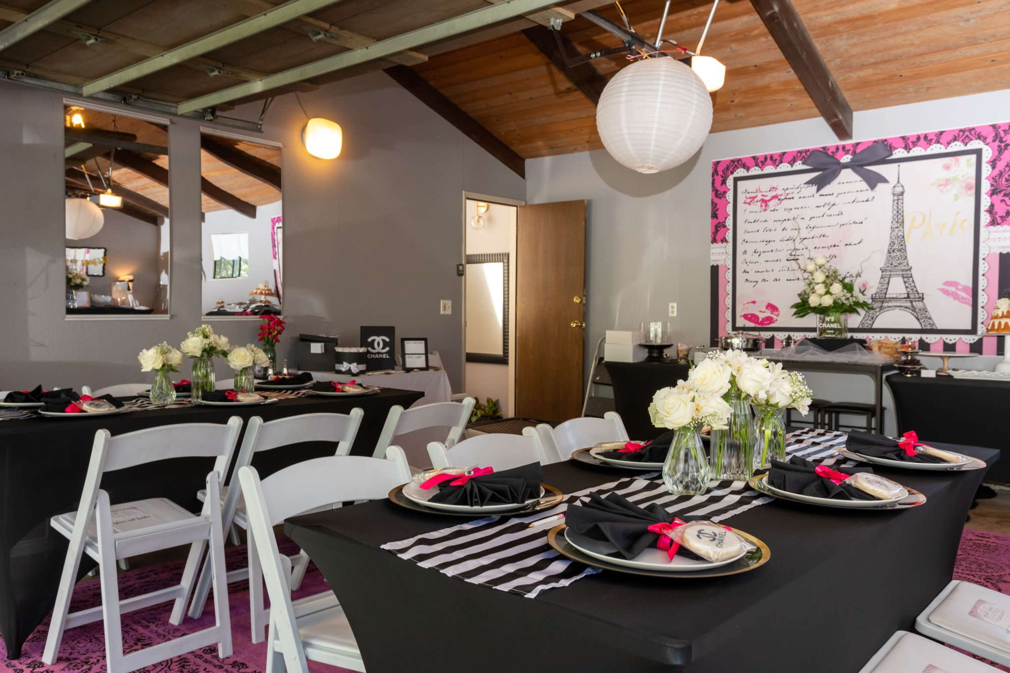 A dining area is set with black tables, white chairs, and floral centerpieces, featuring Parisian-themed decor on the walls.