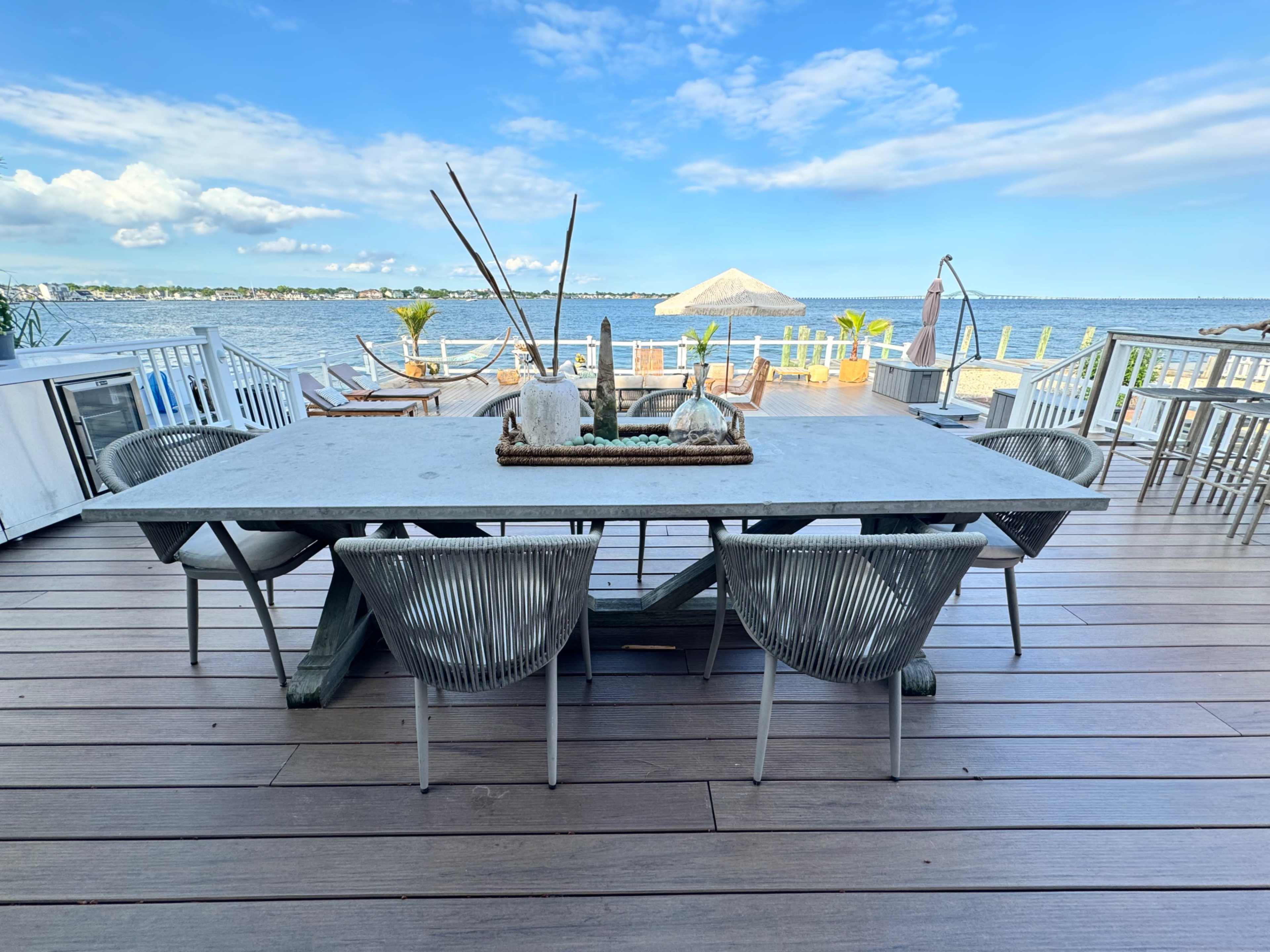A large table with several chairs overlooks a waterfront scene under a clear blue sky.