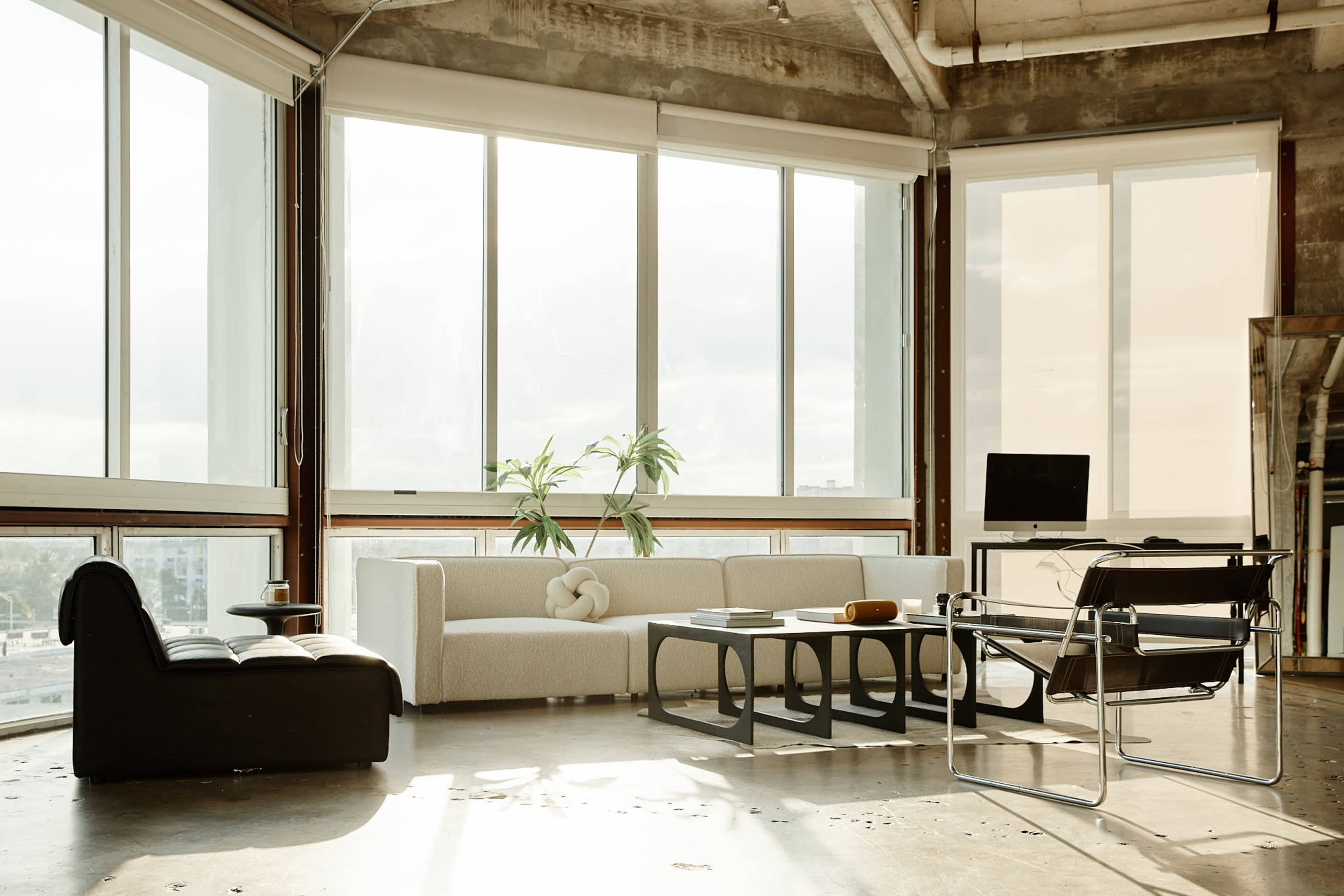 A modern living space with large windows, featuring a light-colored sofa, a black chair, a coffee table, and a desk with a computer.