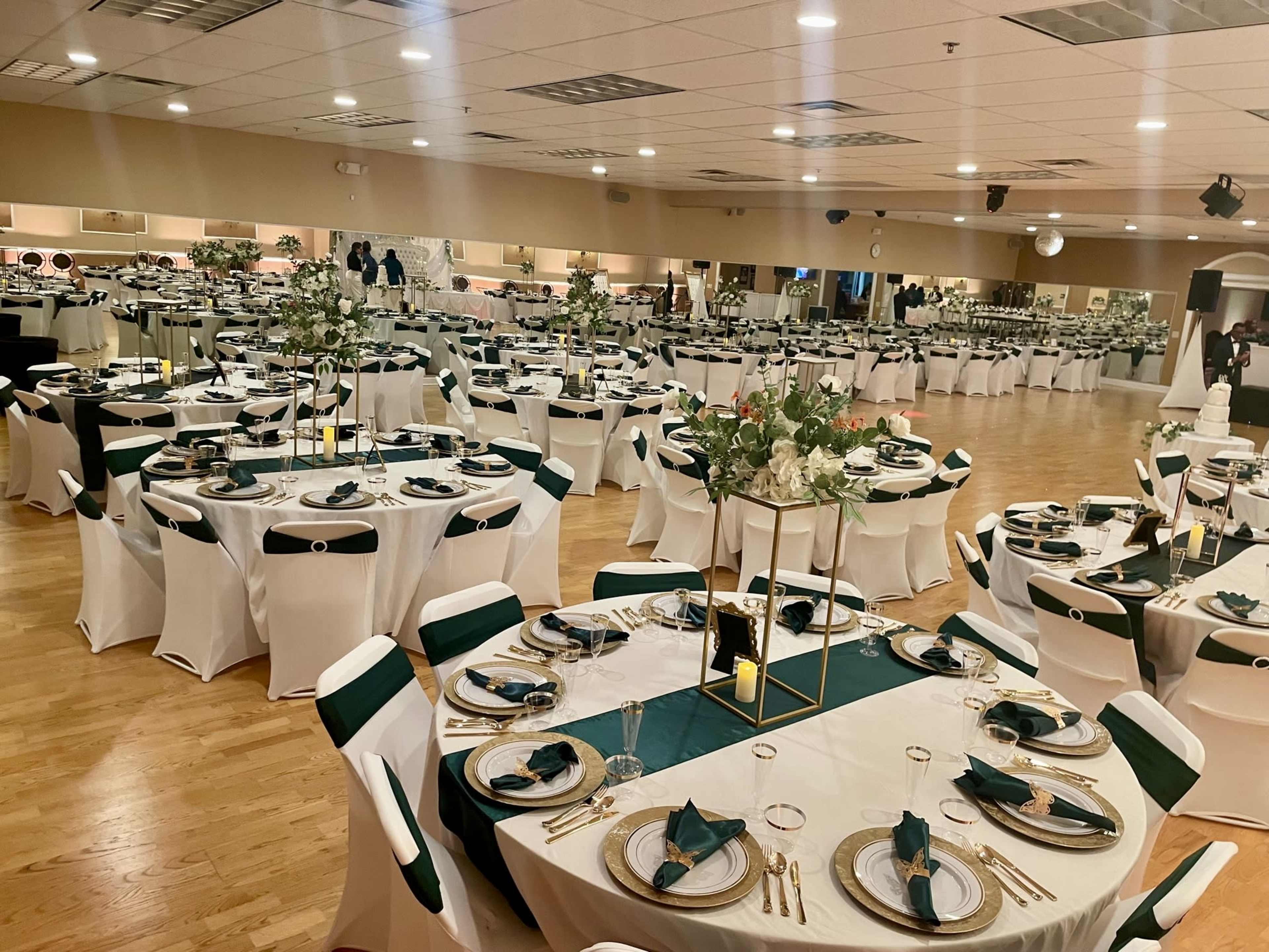 The image shows a banquet hall set up for an event, featuring multiple tables decorated with white tablecloths, dark green accents, and floral centerpieces.