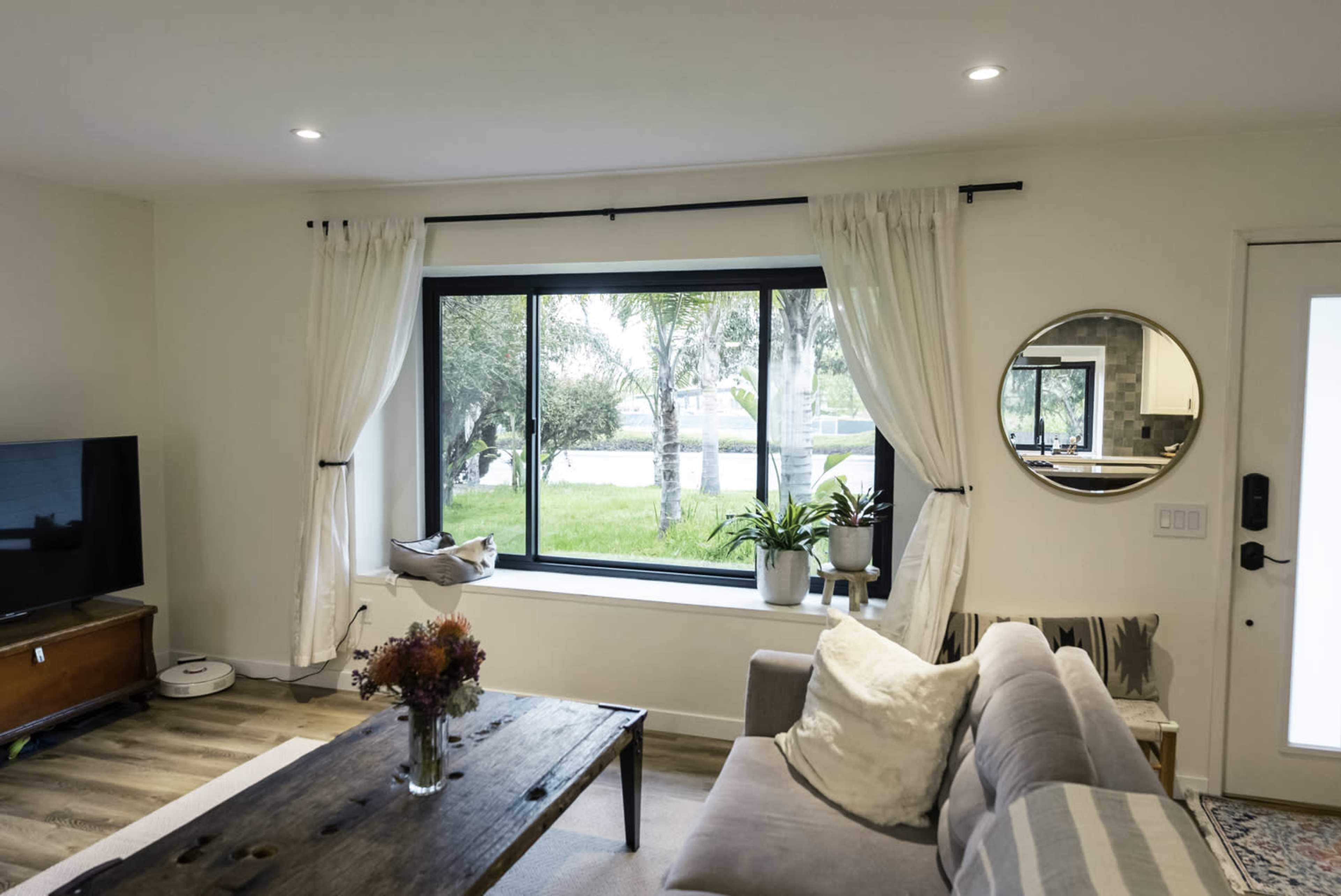 A living room features a large window overlooking a green yard, with a coffee table, sofa, and television present in the space.