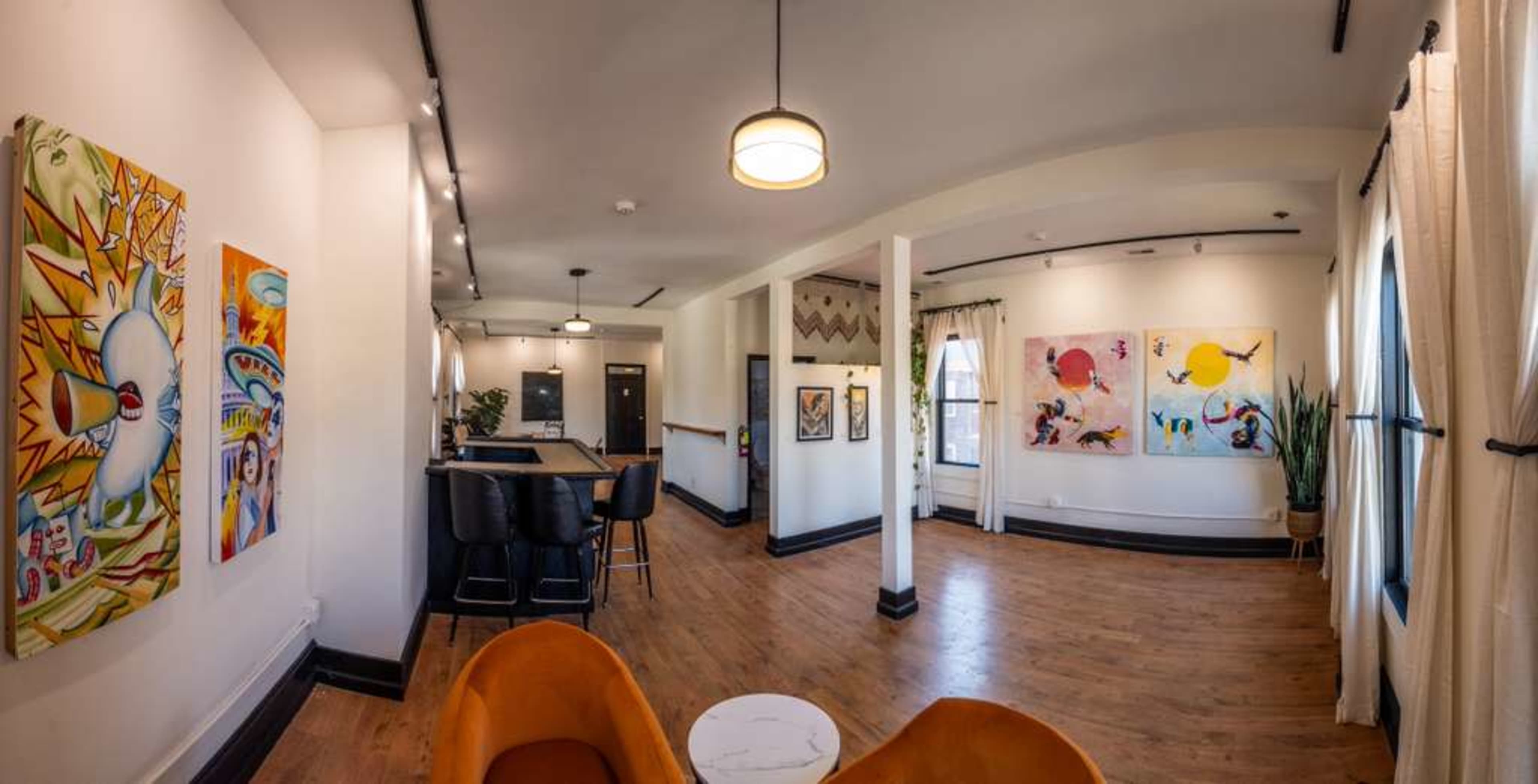 The image shows a spacious interior with hardwood floors, colorful artwork on the walls, and modern furniture including orange chairs and a bar area.