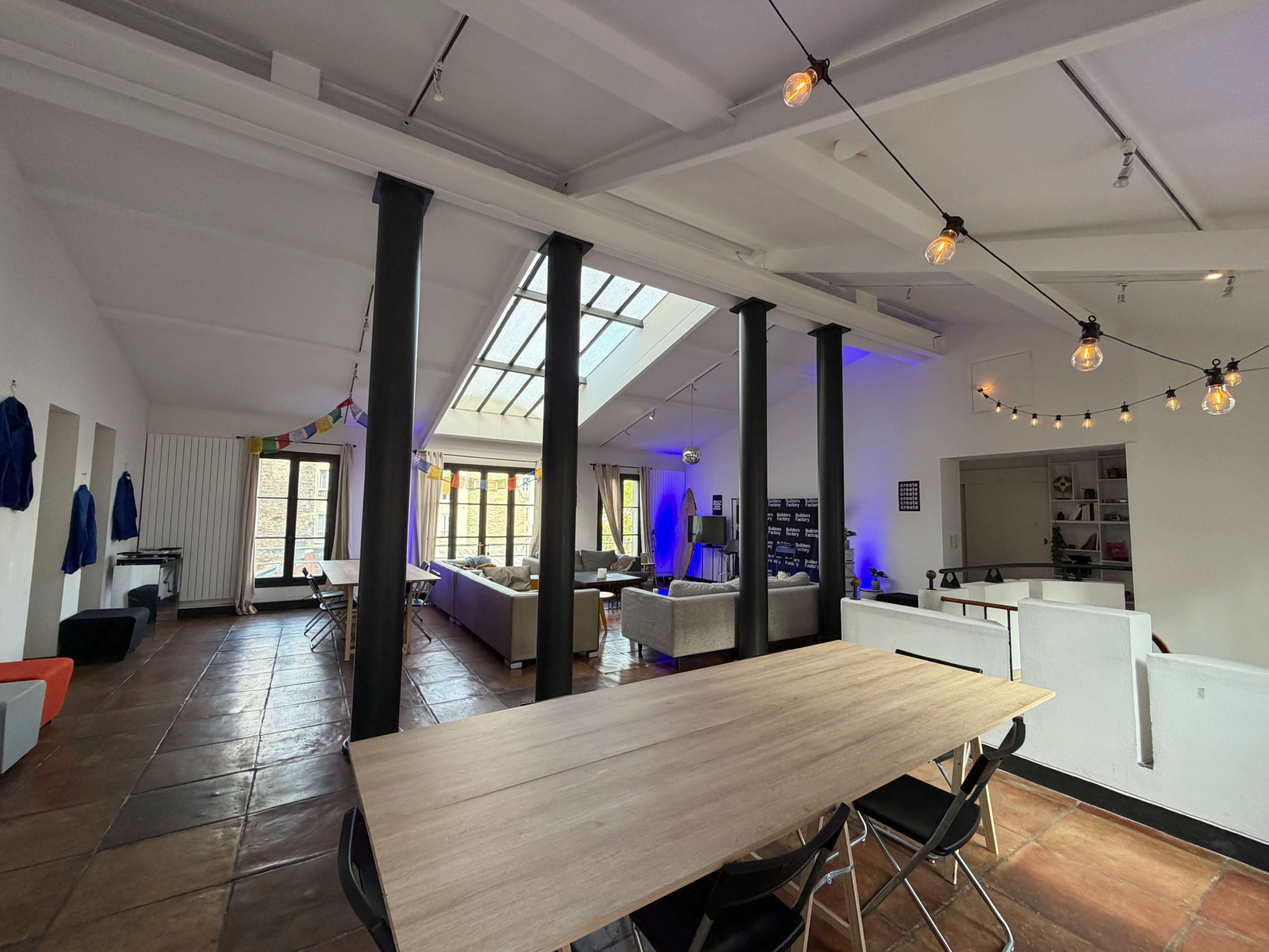 The image shows a spacious, open-plan interior with exposed wooden beams, a large wooden table in the foreground, and various seating areas illuminated by string lights.