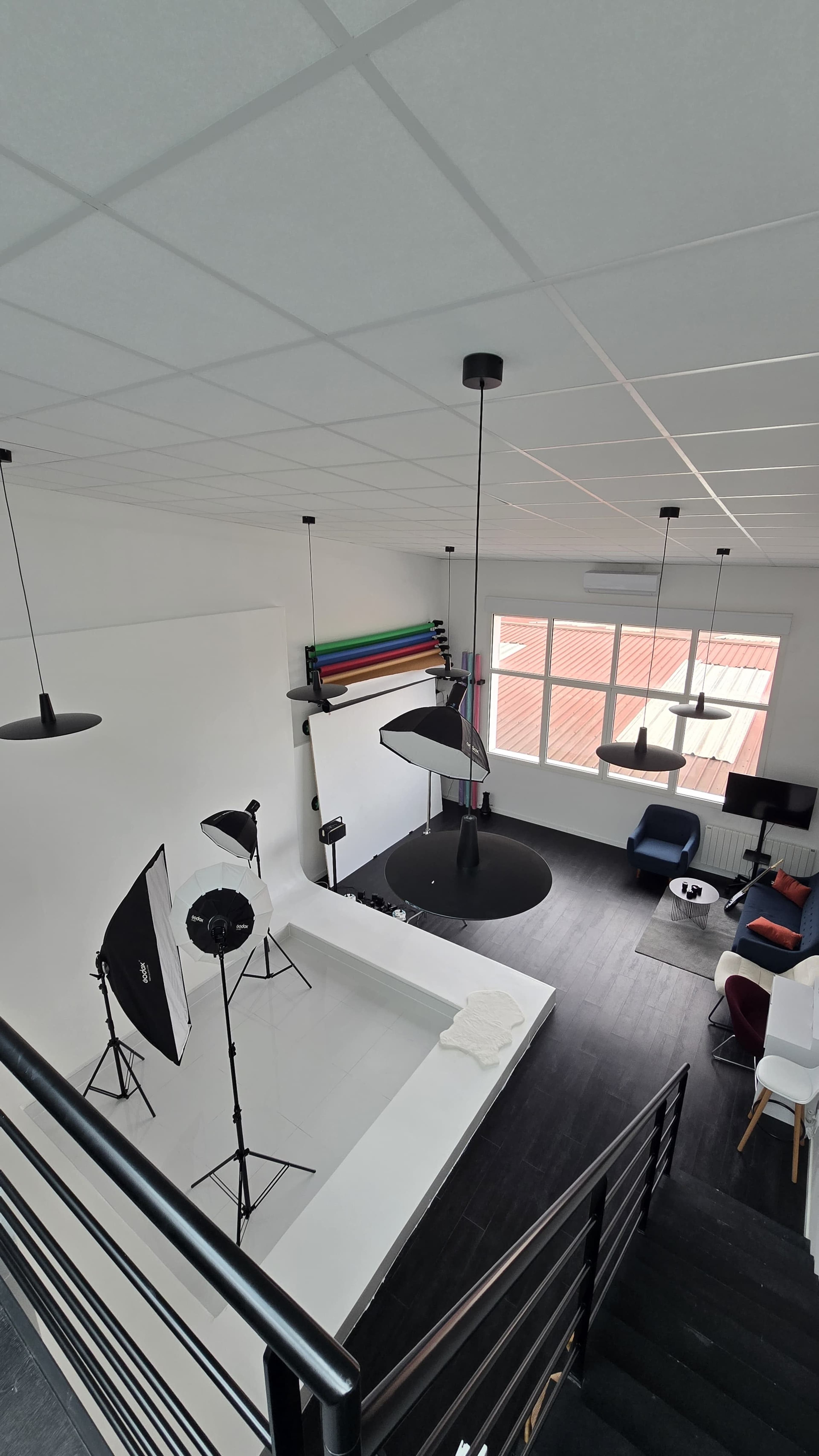 The image shows a photography studio with a light setup, a backdrop, and a seating area, all arranged in a spacious room with large windows.