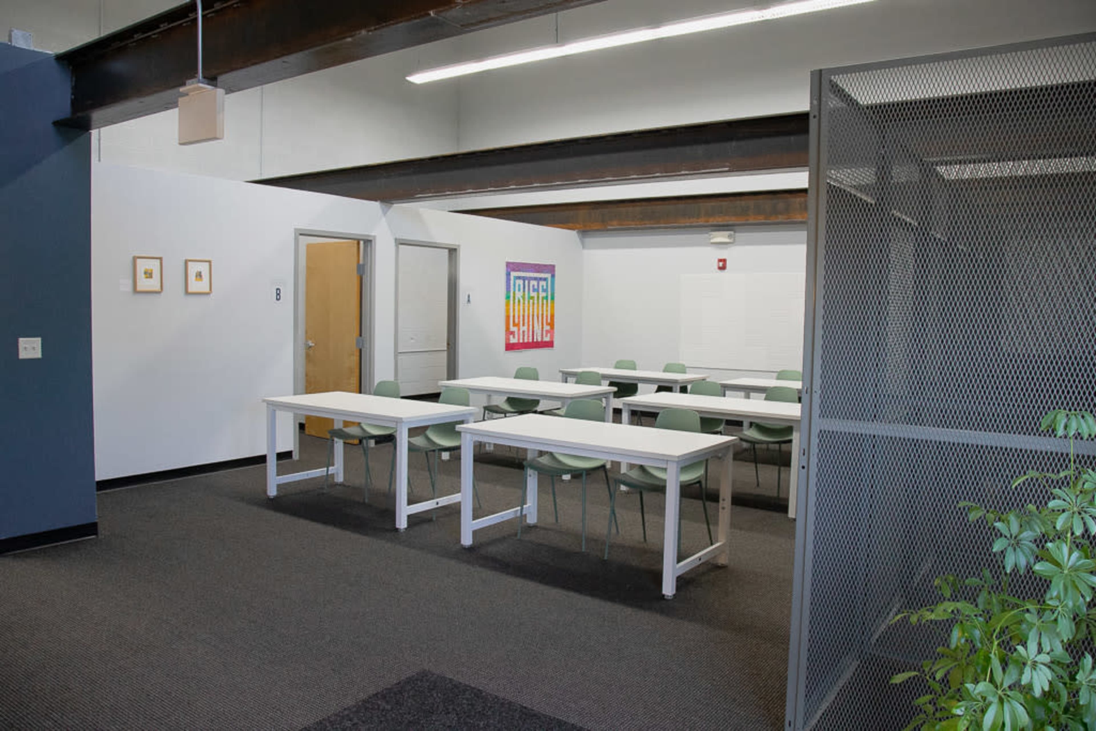 A classroom with several white tables and green chairs arranged in neat rows, and a colorful artwork displayed on the wall.