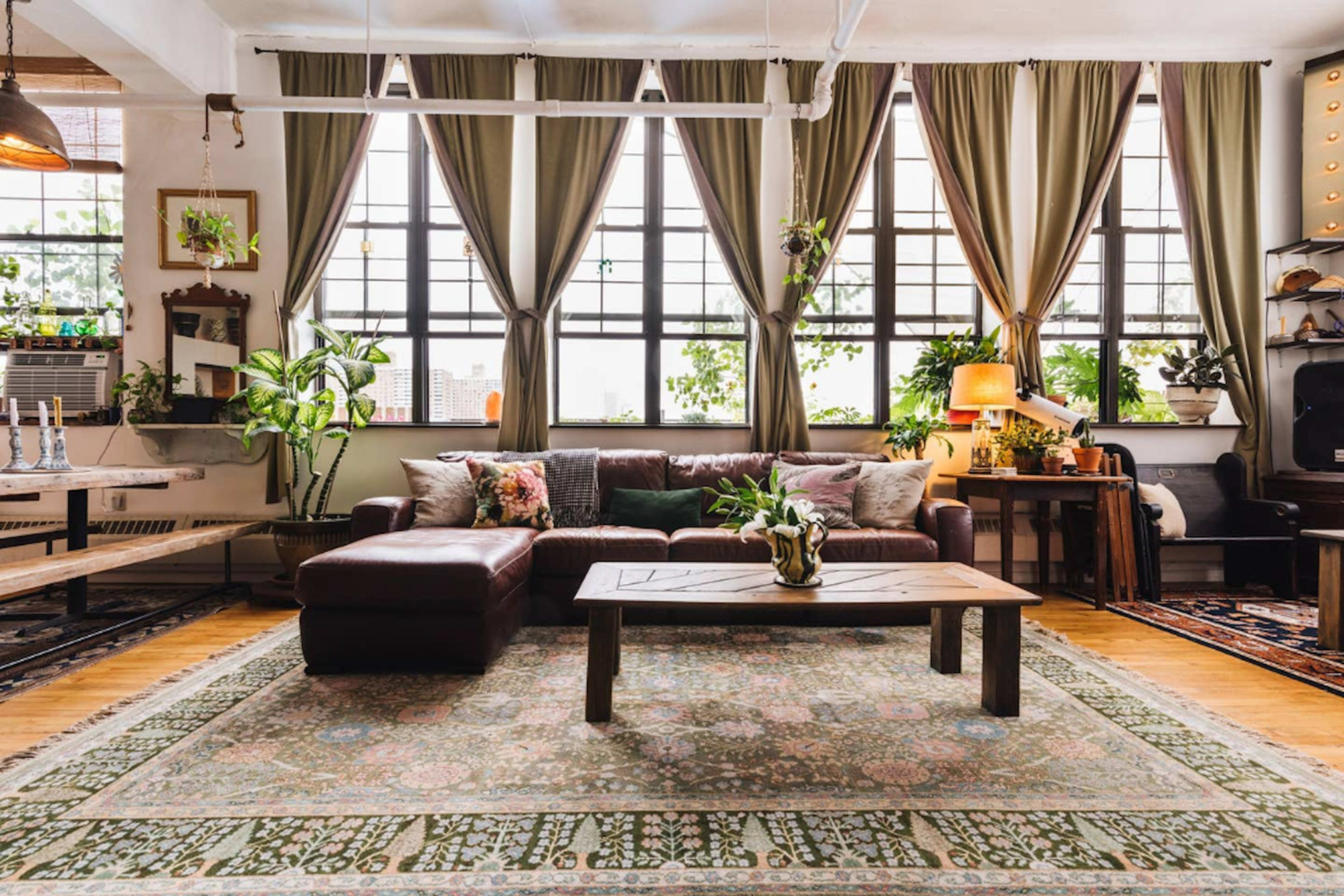The image shows a cozy living room with large windows, a brown sectional couch, a wooden coffee table, and various plants arranged around the space.
