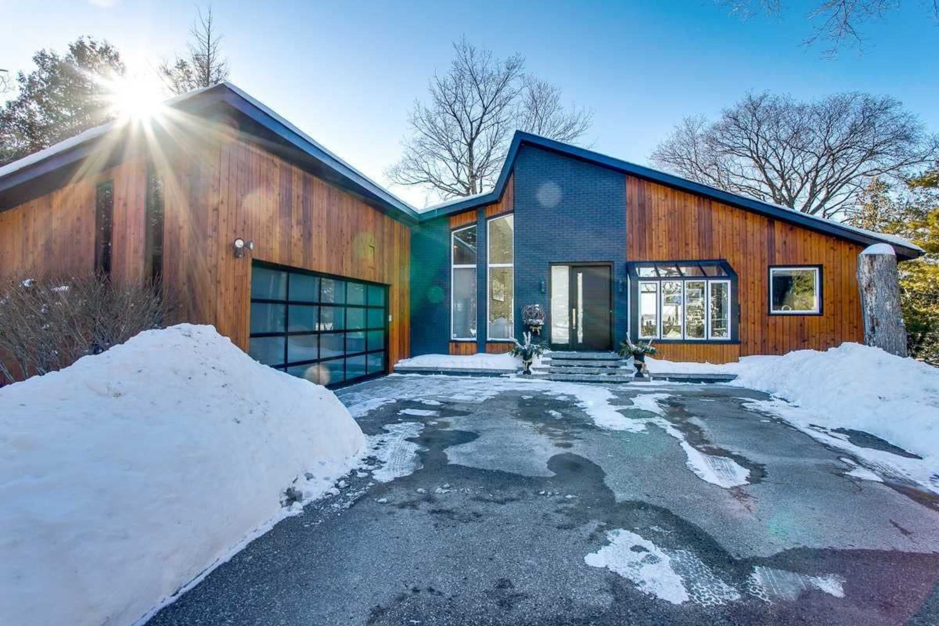 A modern house with wooden siding and a large garage sits on a snowy driveway under clear blue skies.