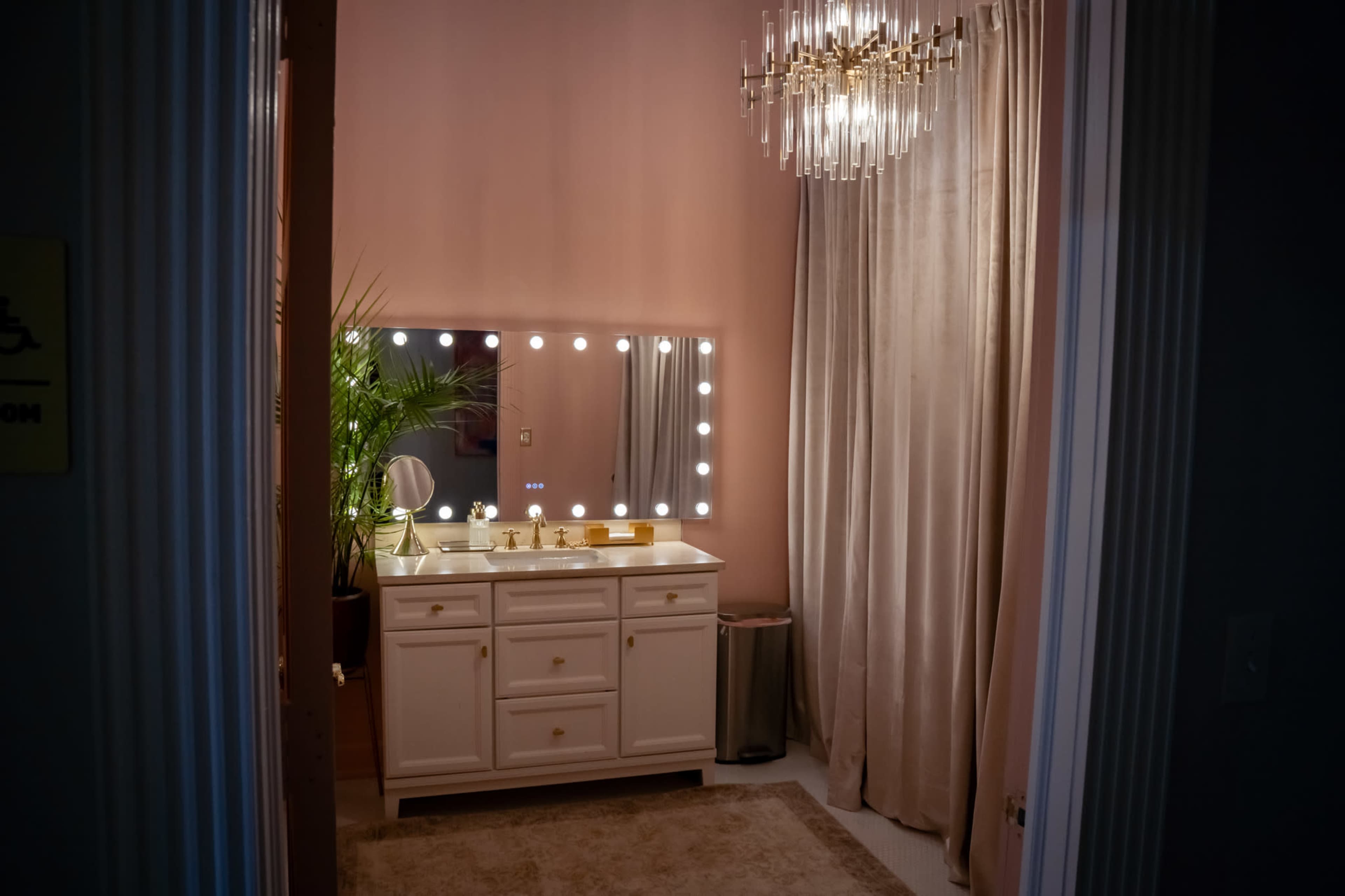The image shows a well-lit bathroom with a vanity area featuring a large mirror framed by light bulbs, a small plant, and curtains on one side.