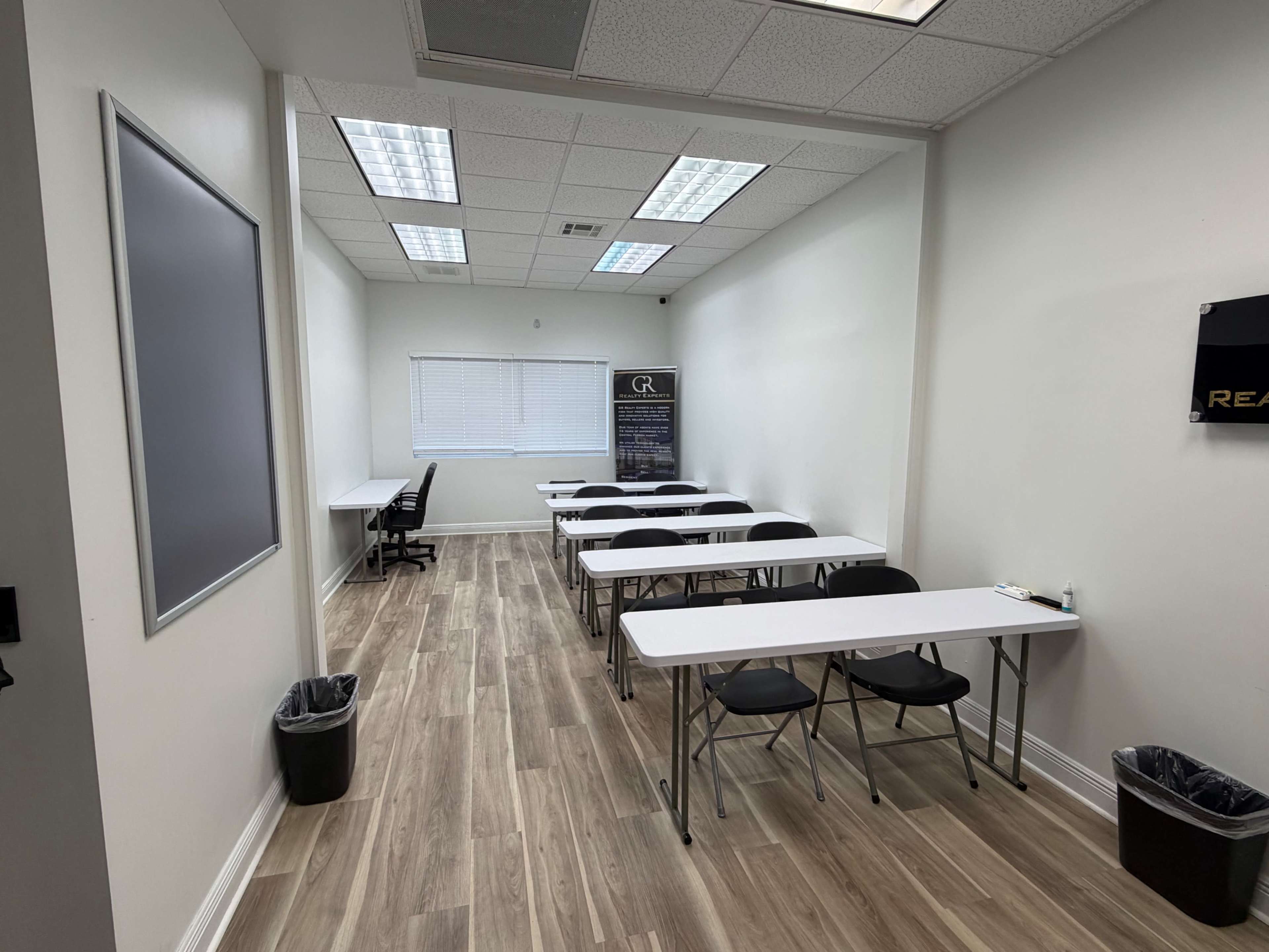 The image shows a classroom with several empty folding tables and chairs arranged in rows, complemented by a window that allows natural light to enter.