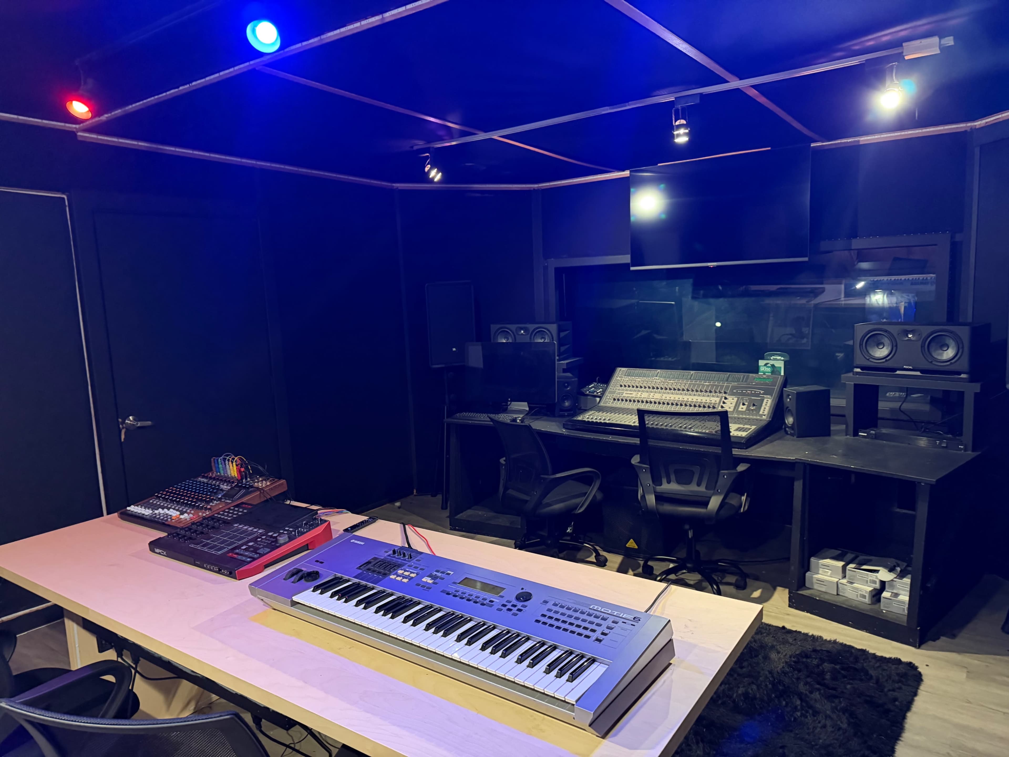 The image shows a music production studio featuring a keyboard on a desk, a mixing console at the back, and studio monitors on either side, illuminated by colored lights on the ceiling.