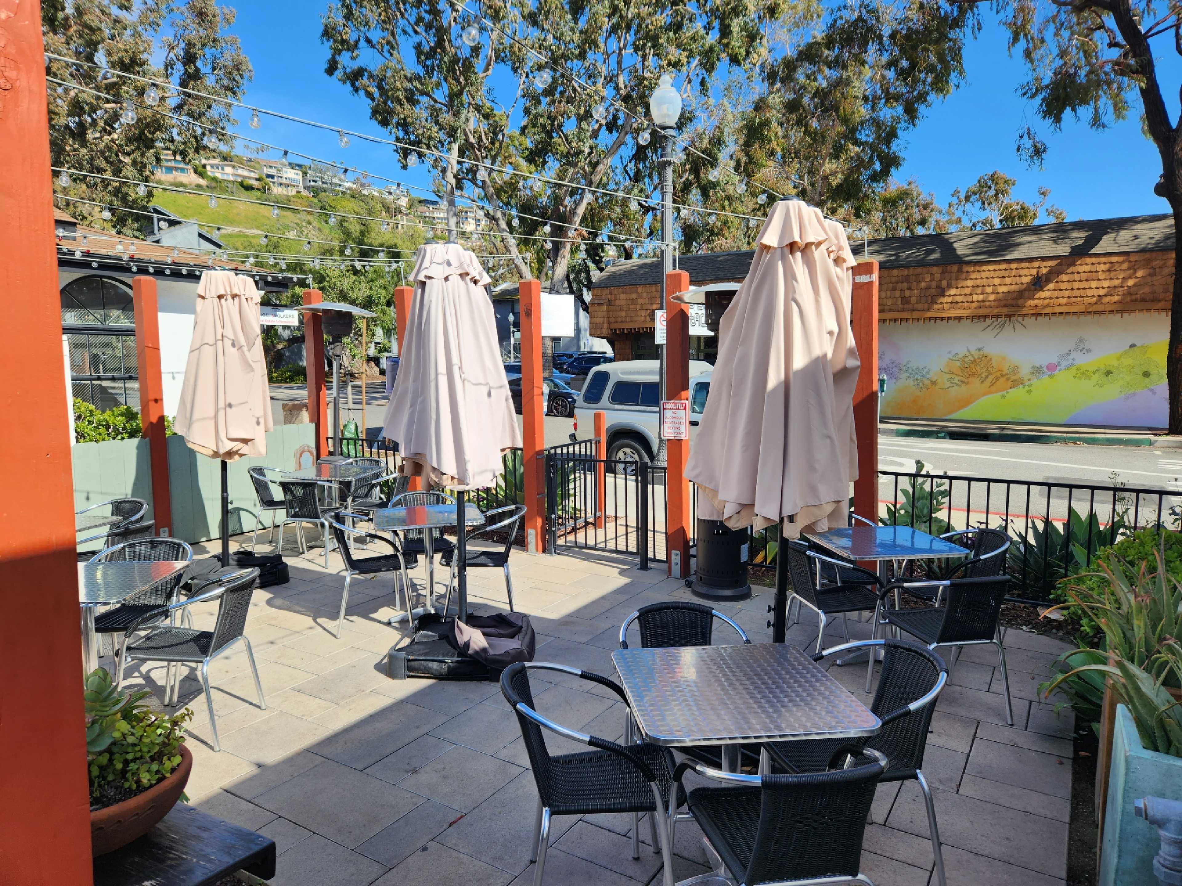 A patio area with empty tables and umbrellas is visible, surrounded by greenery and a street view in the background.
