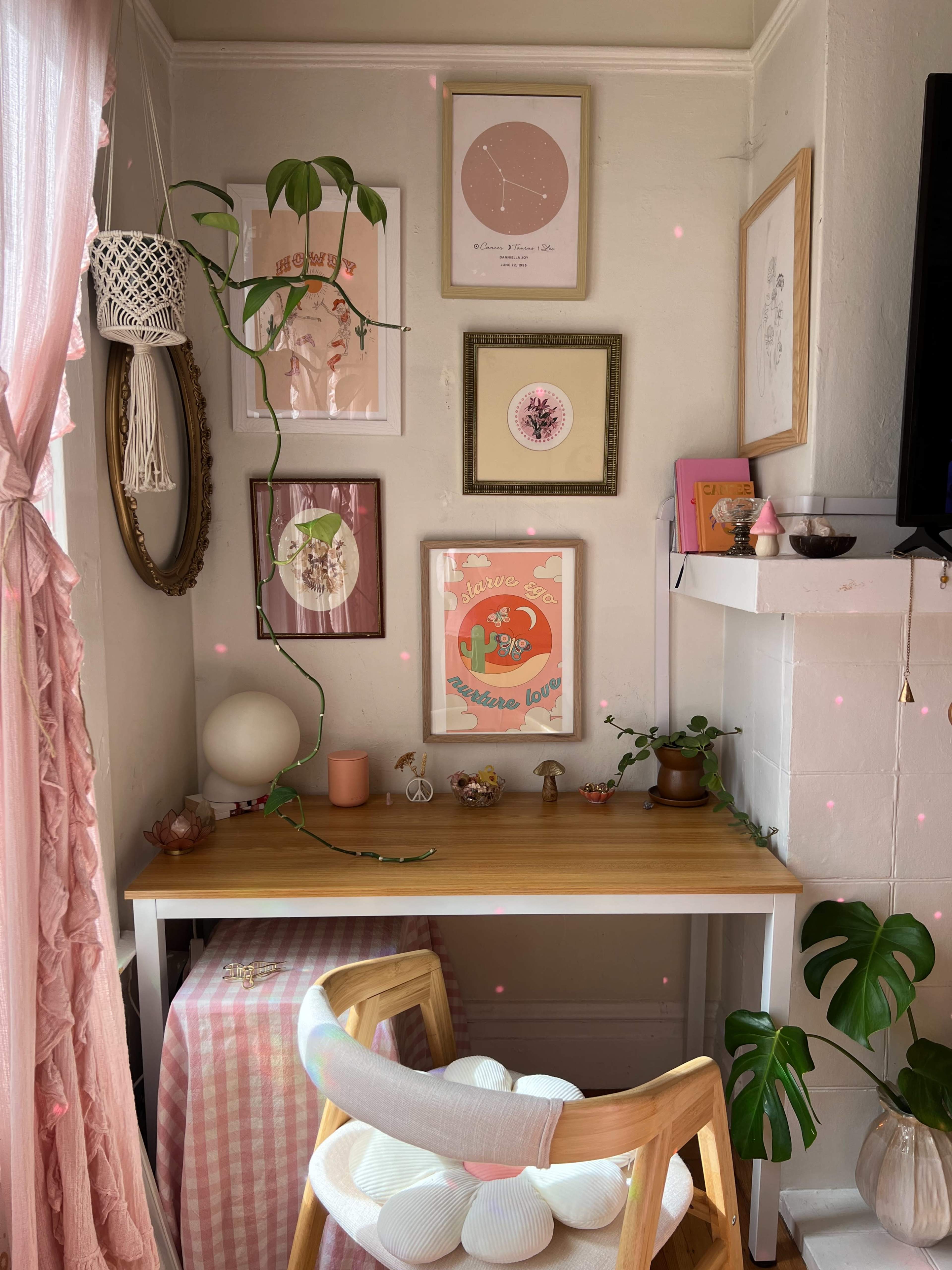 A small workspace features a wooden desk with decorative art pieces on the wall, a plant climbing, and a pink tablecloth draped on a nearby chair.